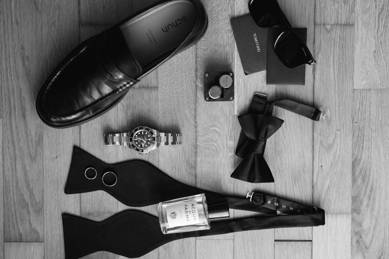 Flat lay of black formal accessories and shoes arranged on wooden floor.