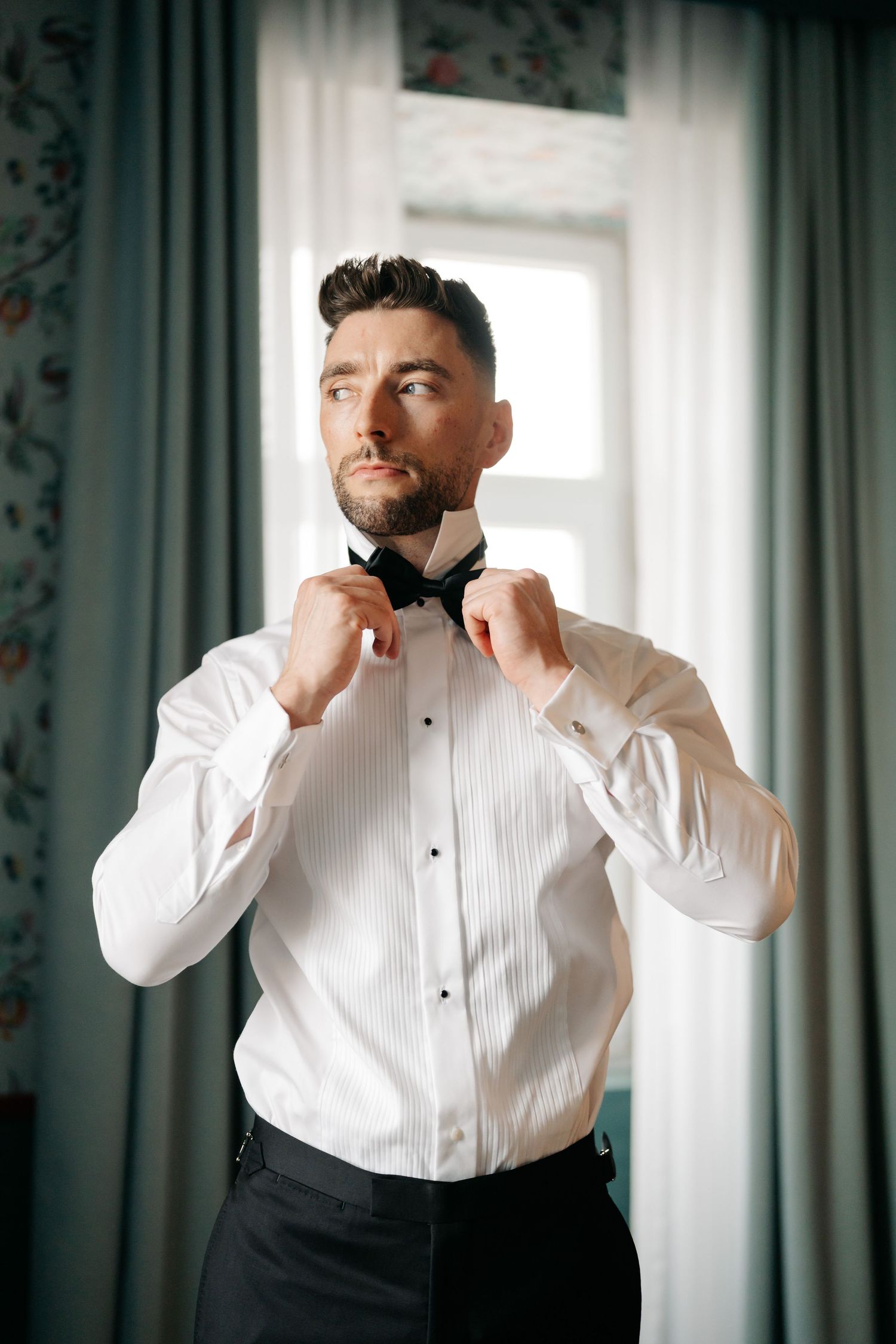 Person in white dress shirt adjusts collar near window with floral wallpaper.