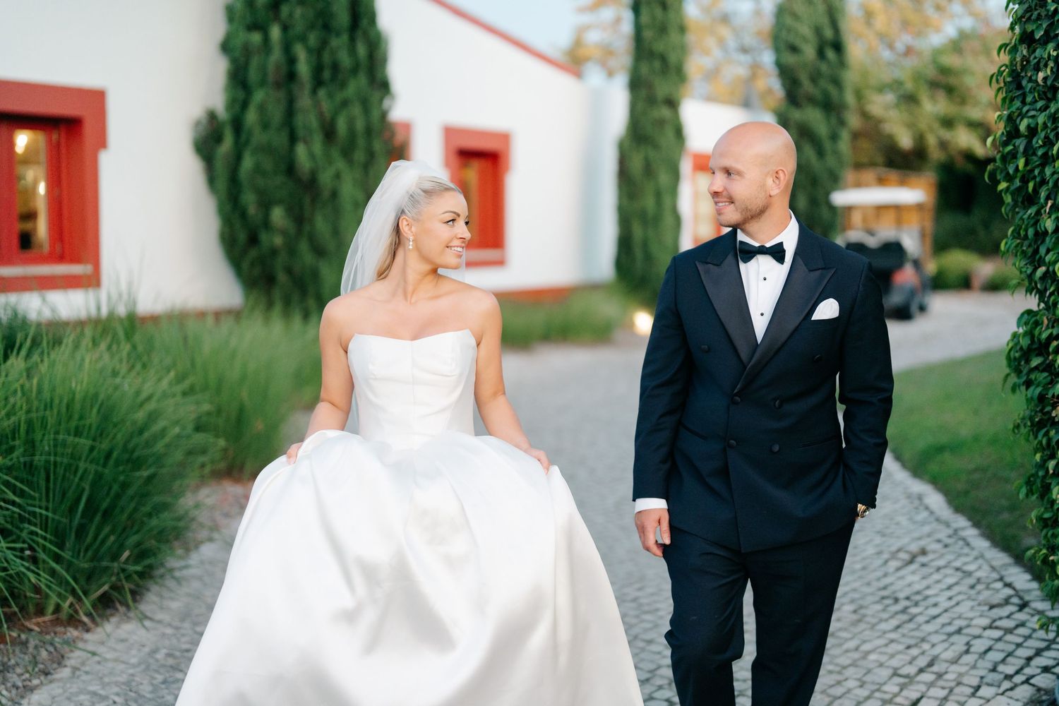 Elegant wedding couple poses together on a stone path surrounded by lush greenery and red-trimmed architecture.