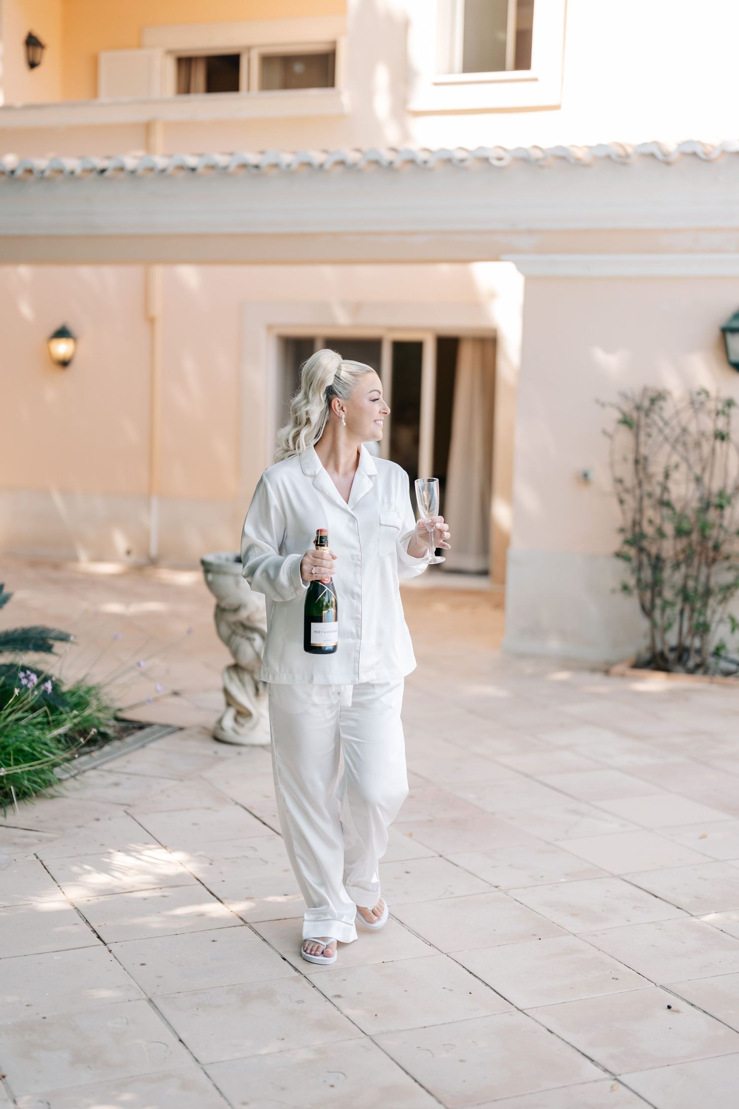 Figure in white outfit walking on stone patio with champagne.