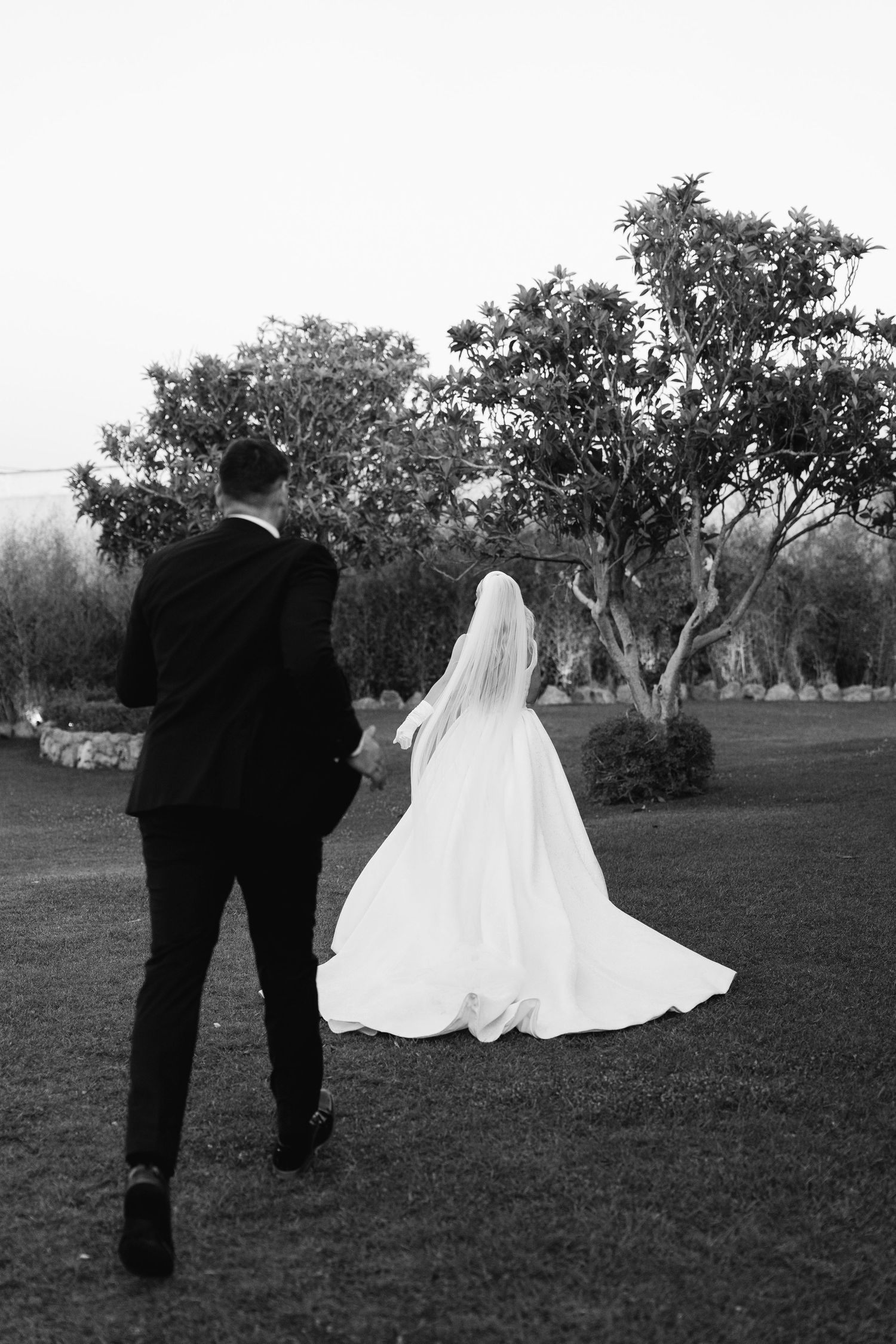 A couple in wedding attire walks across a grassy lawn with trees in the background.