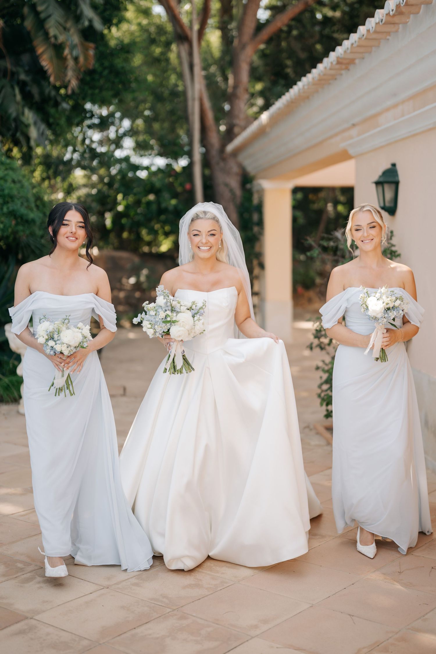 Bridesmaids in elegant white dresses sitting on stone steps during an outdoor wedding celebration.
