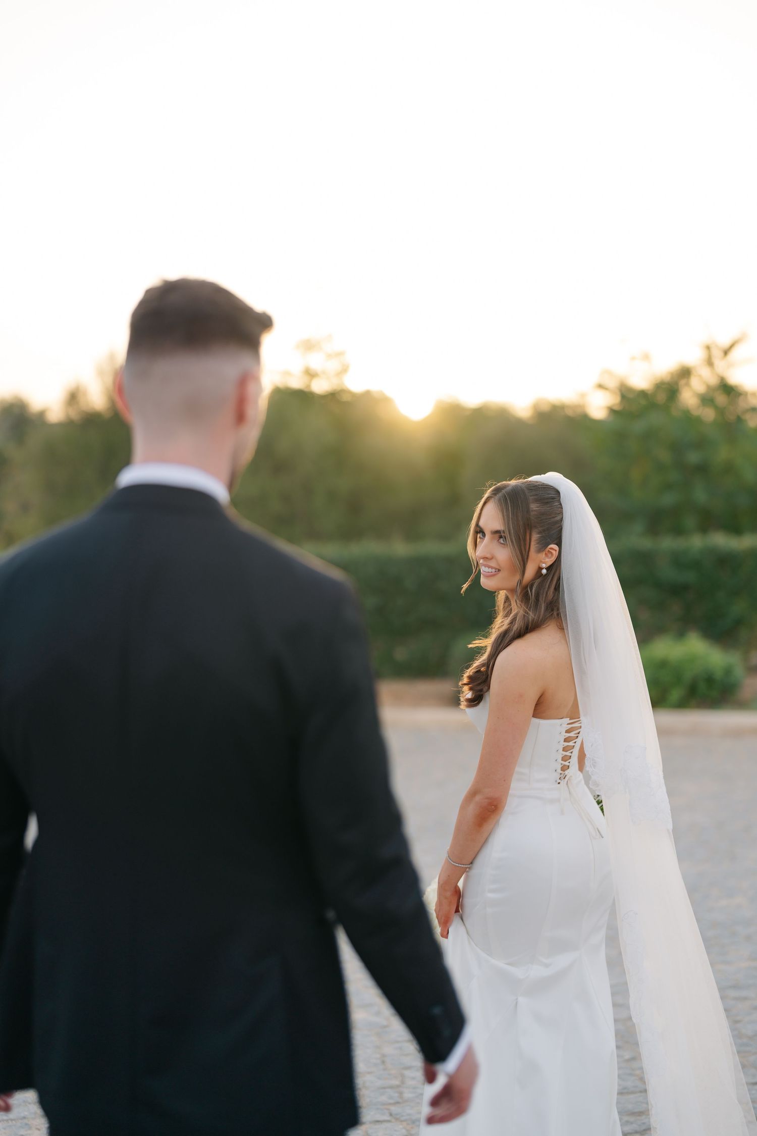 A bride in a fitted white dress looks back at her groom during golden hour.