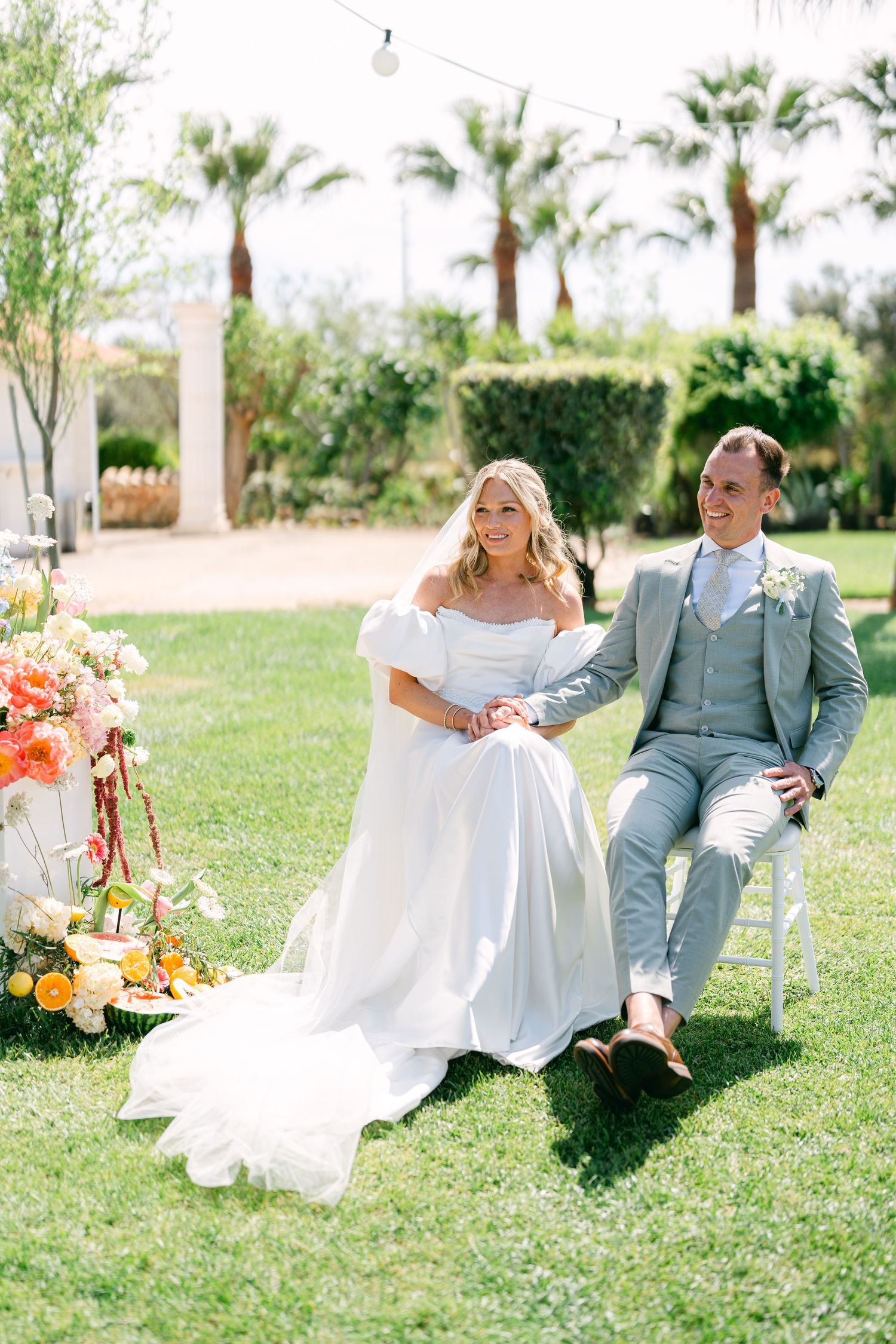 Wedding couple relaxes on white chairs during outdoor garden photo session with tropical backdrop.