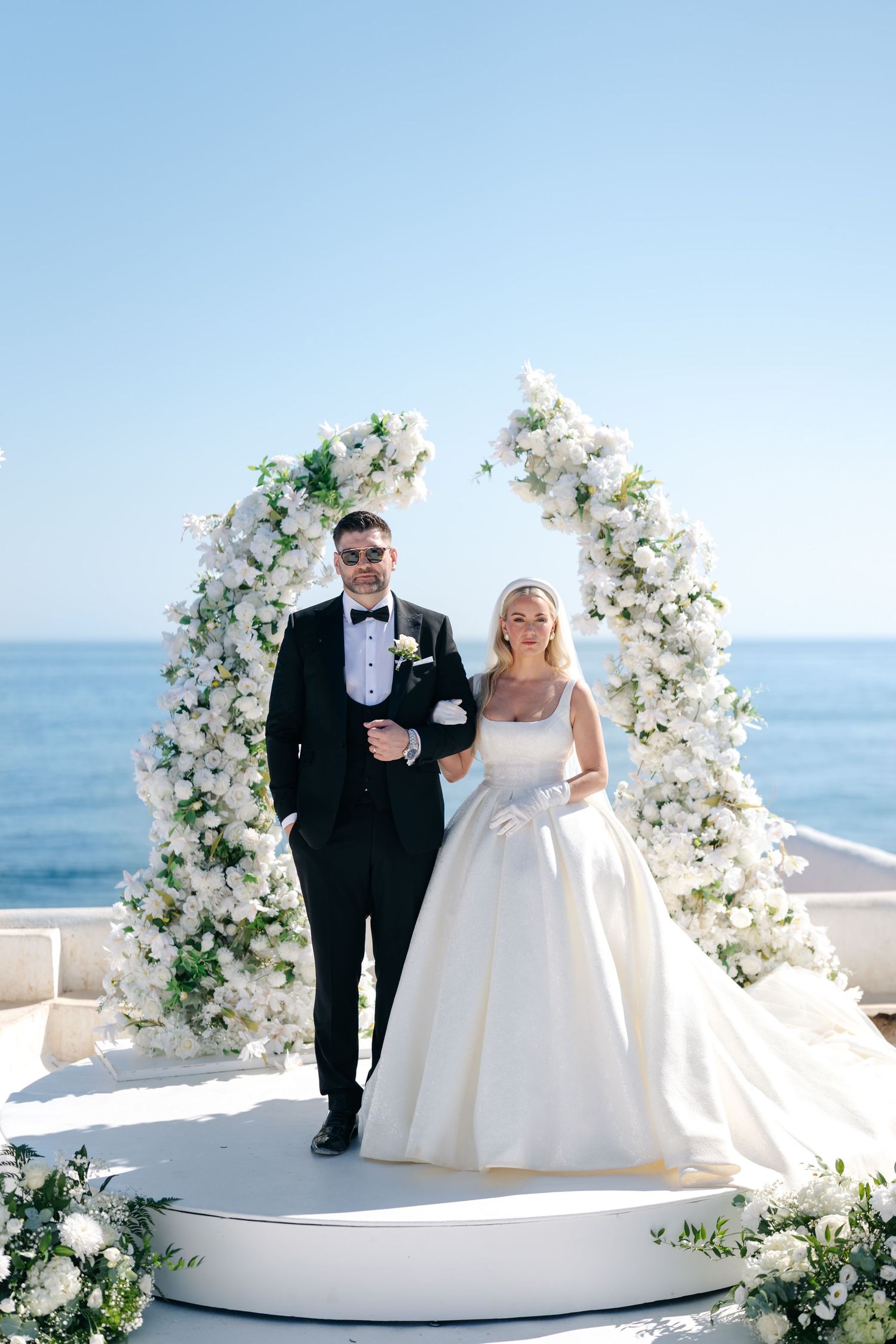 Newlyweds pose on a white platform decorated with floral arrangements by the ocean.