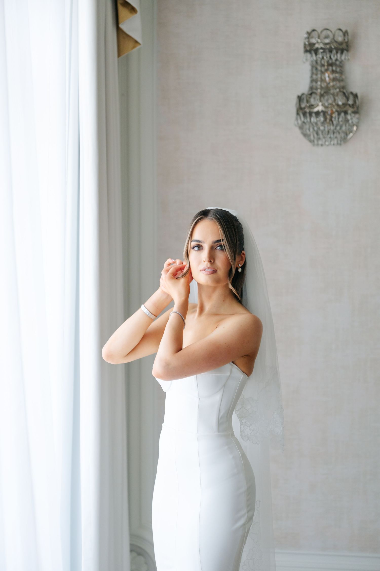 A bride in a fitted white gown stands against a light wall with an ornate chandelier.