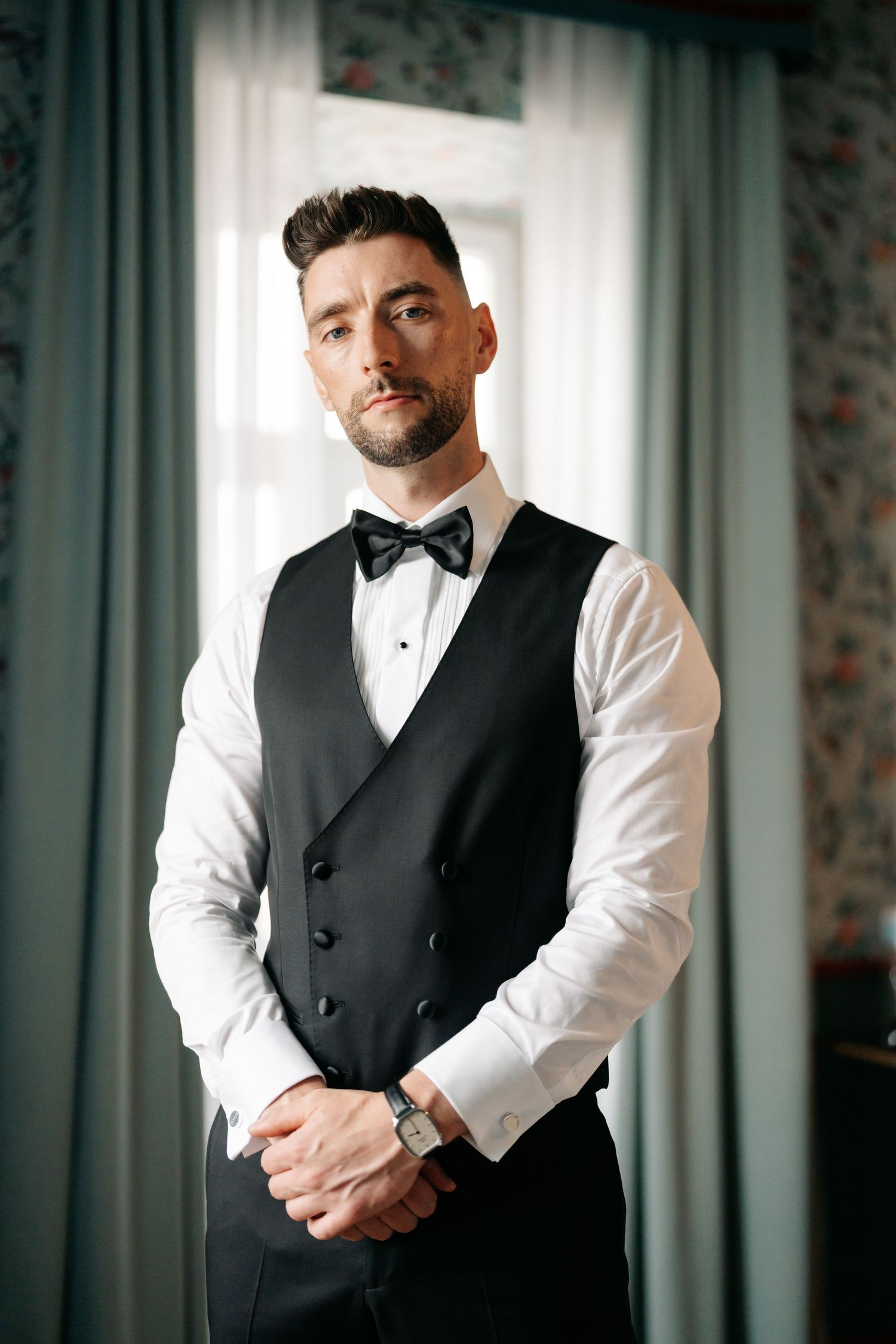 Classic formal wear with double-breasted black vest and bow tie against window light.