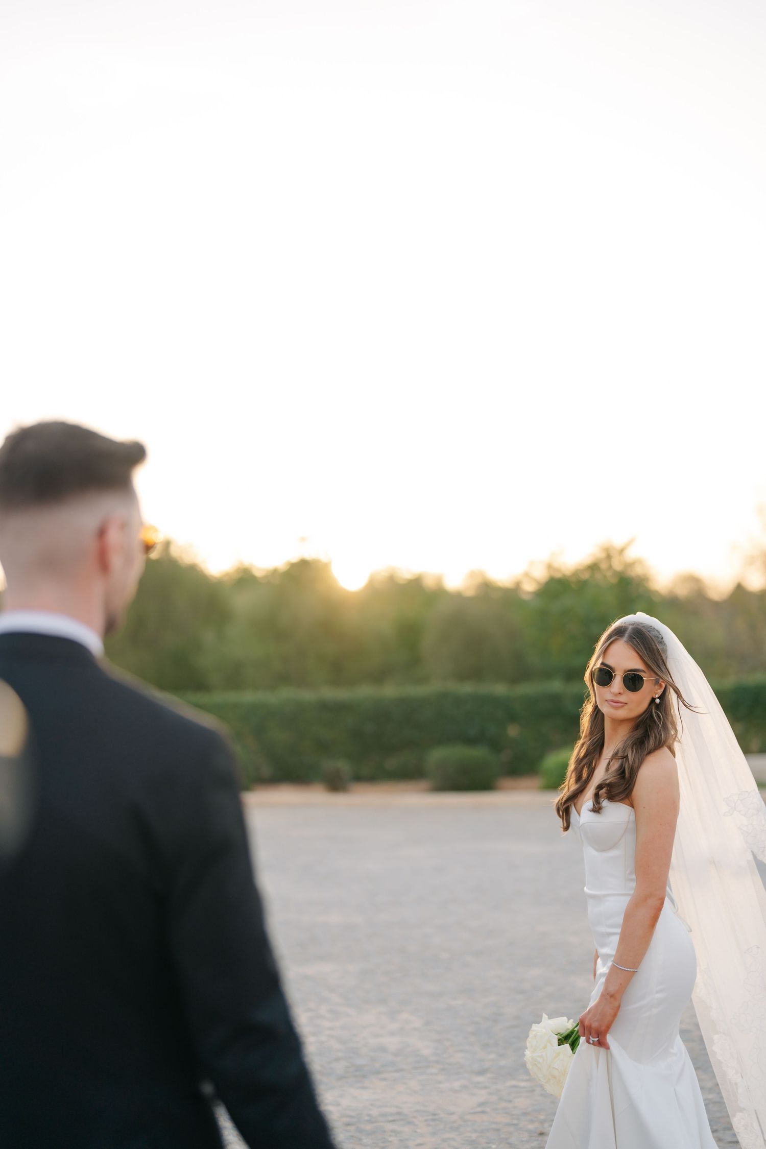 A bride in sunglasses and white dress stands in golden sunset light while looking at her groom.