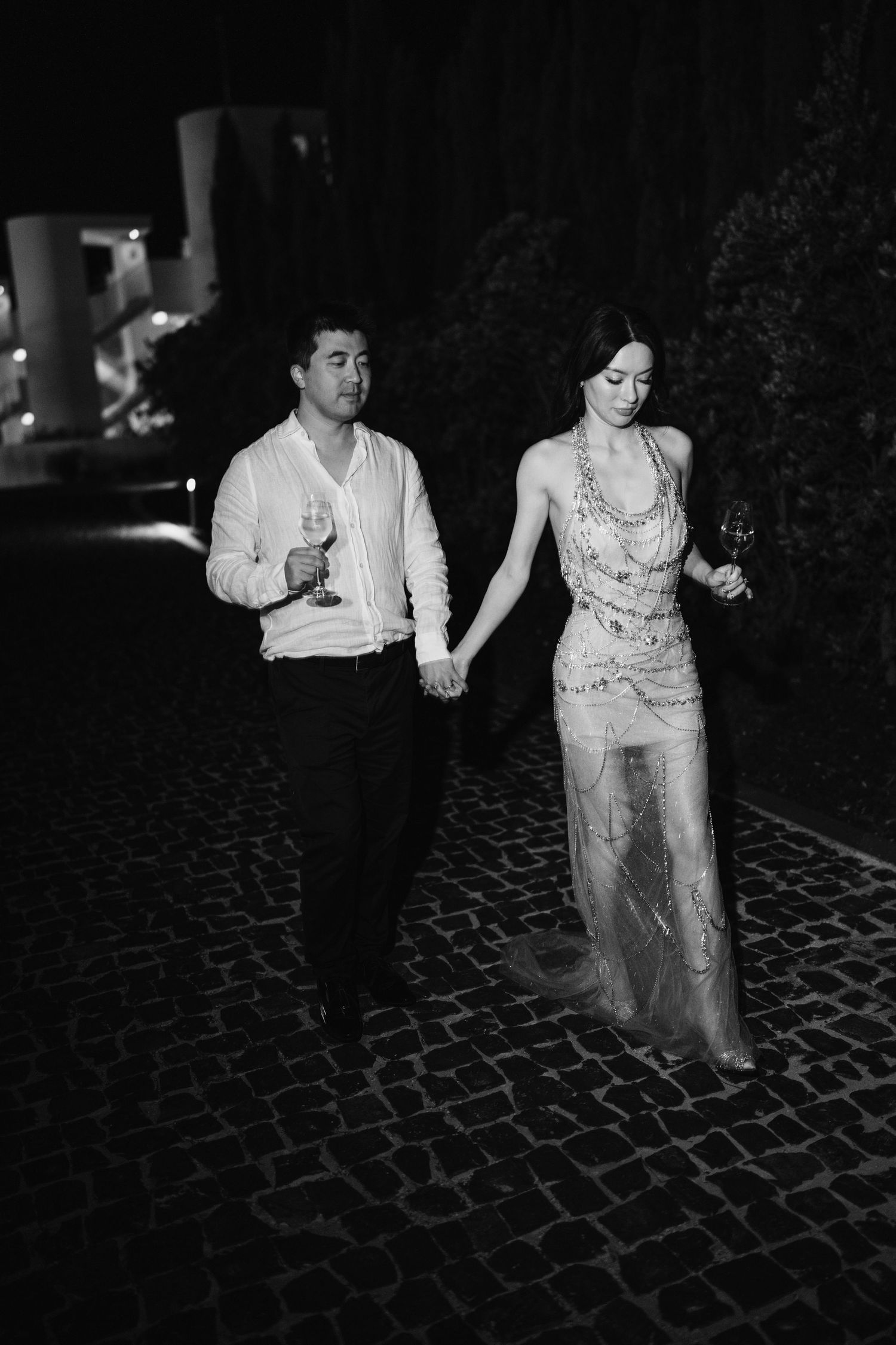 Couple holding hands while walking on cobblestone pavement at night with wine glasses.