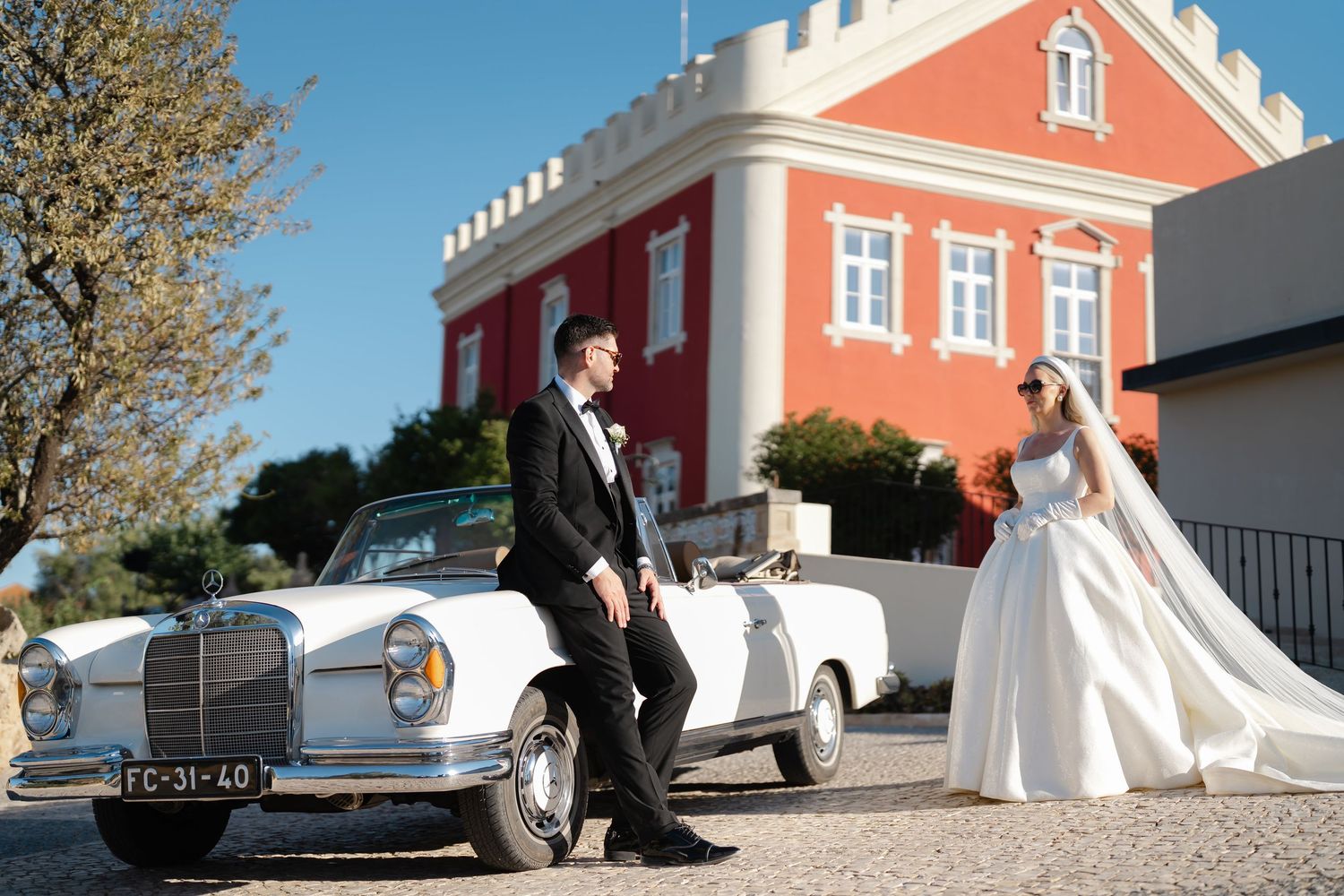 Vintage Mercedes-Benz wedding car parked in front of a striking coral-colored castle building.