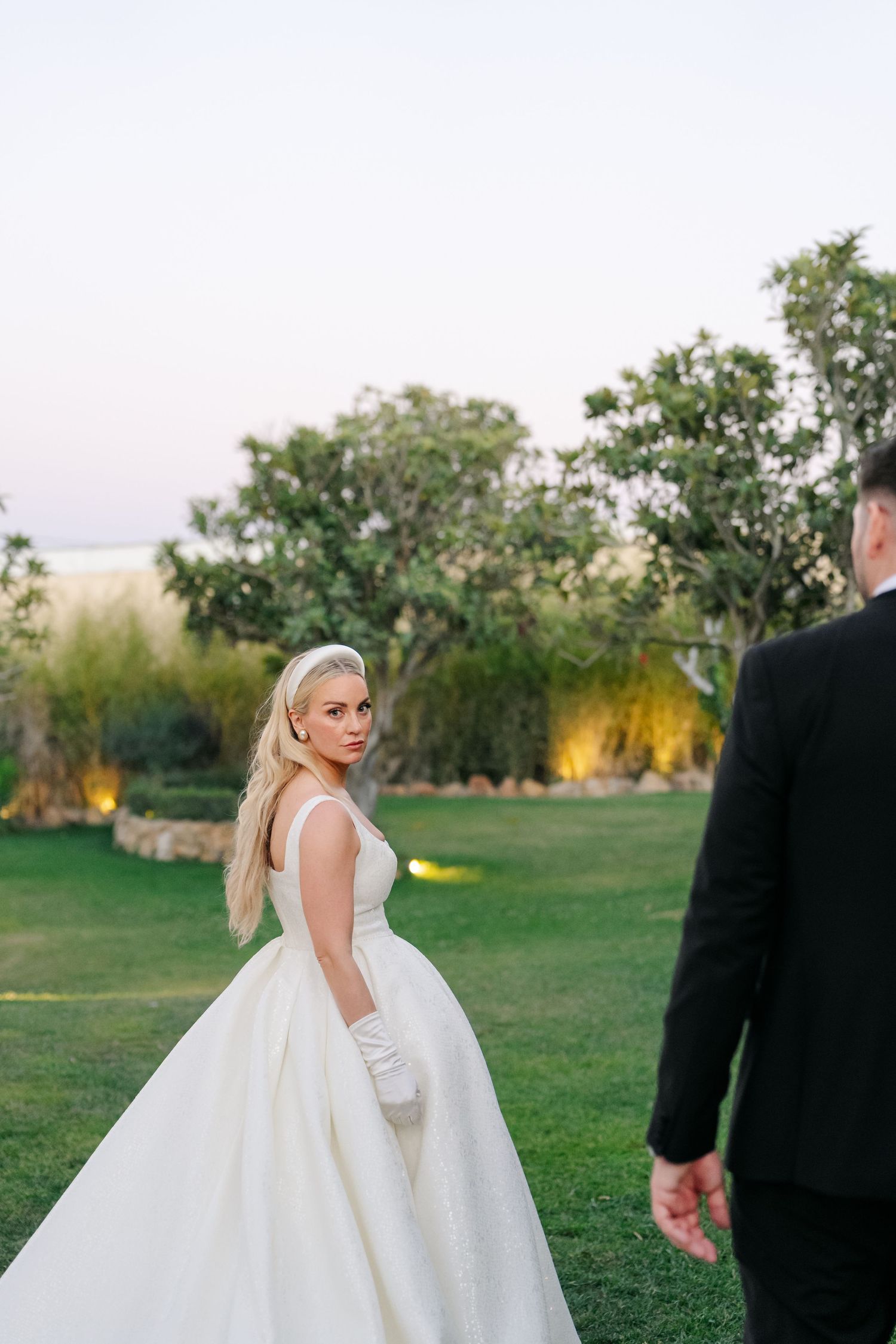 A bride in a flowing white ballgown poses gracefully in an outdoor garden setting at sunset.