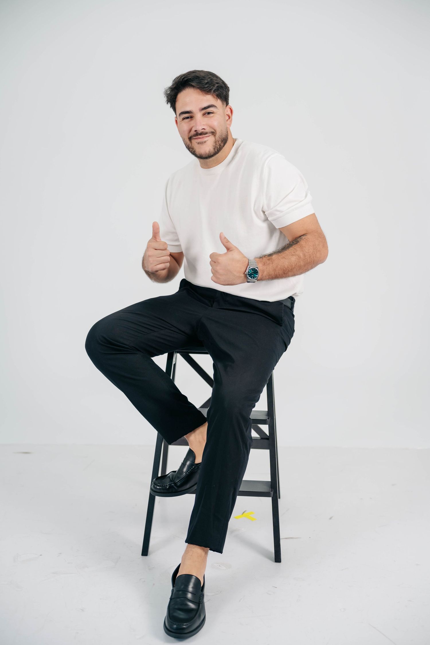 Person in casual white t-shirt and black pants sits on stool giving thumbs up against light background.