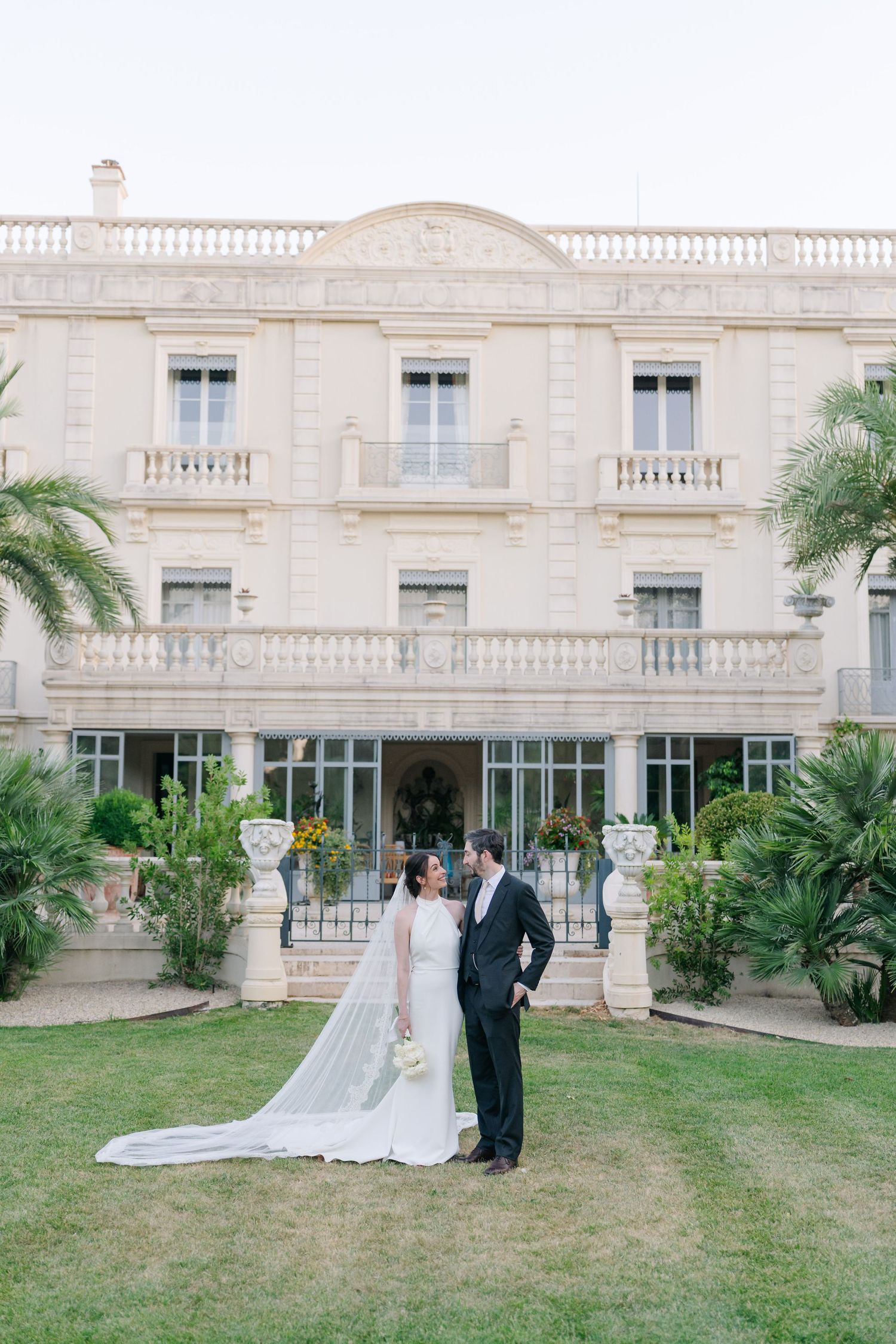 Bride and groom posing on green lawn before elegant white European villa with balconies and tropical palms.