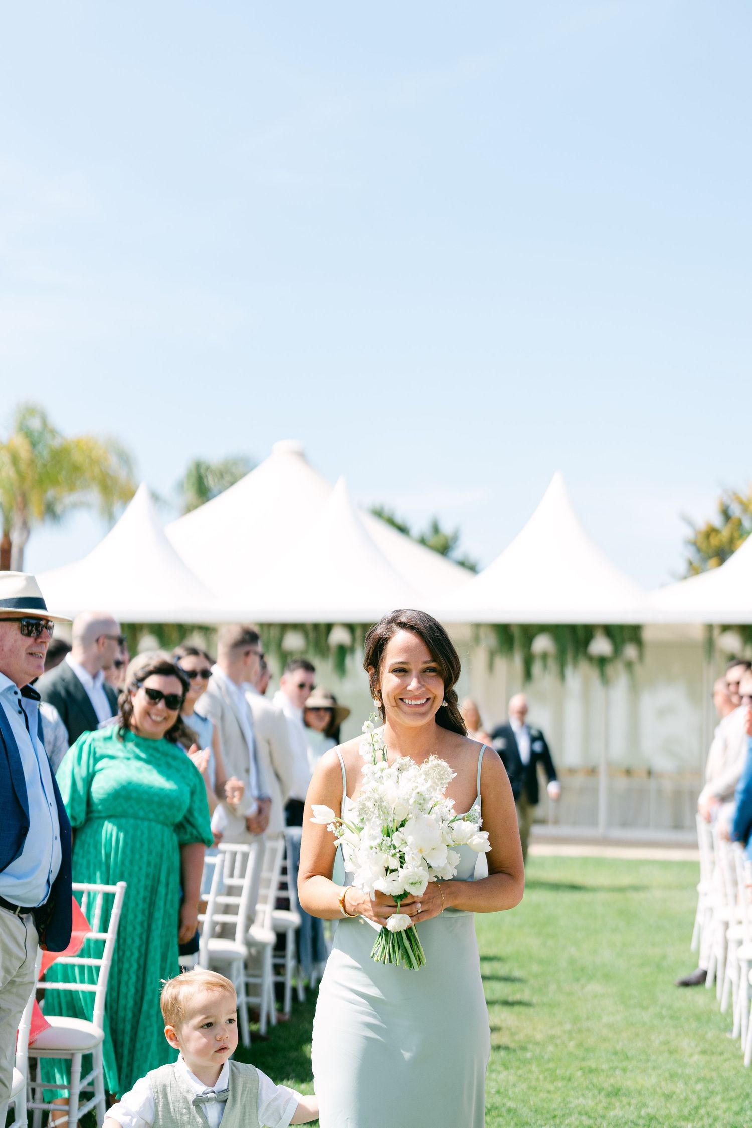 Wedding party members walk down the aisle at an outdoor ceremony with white tents.