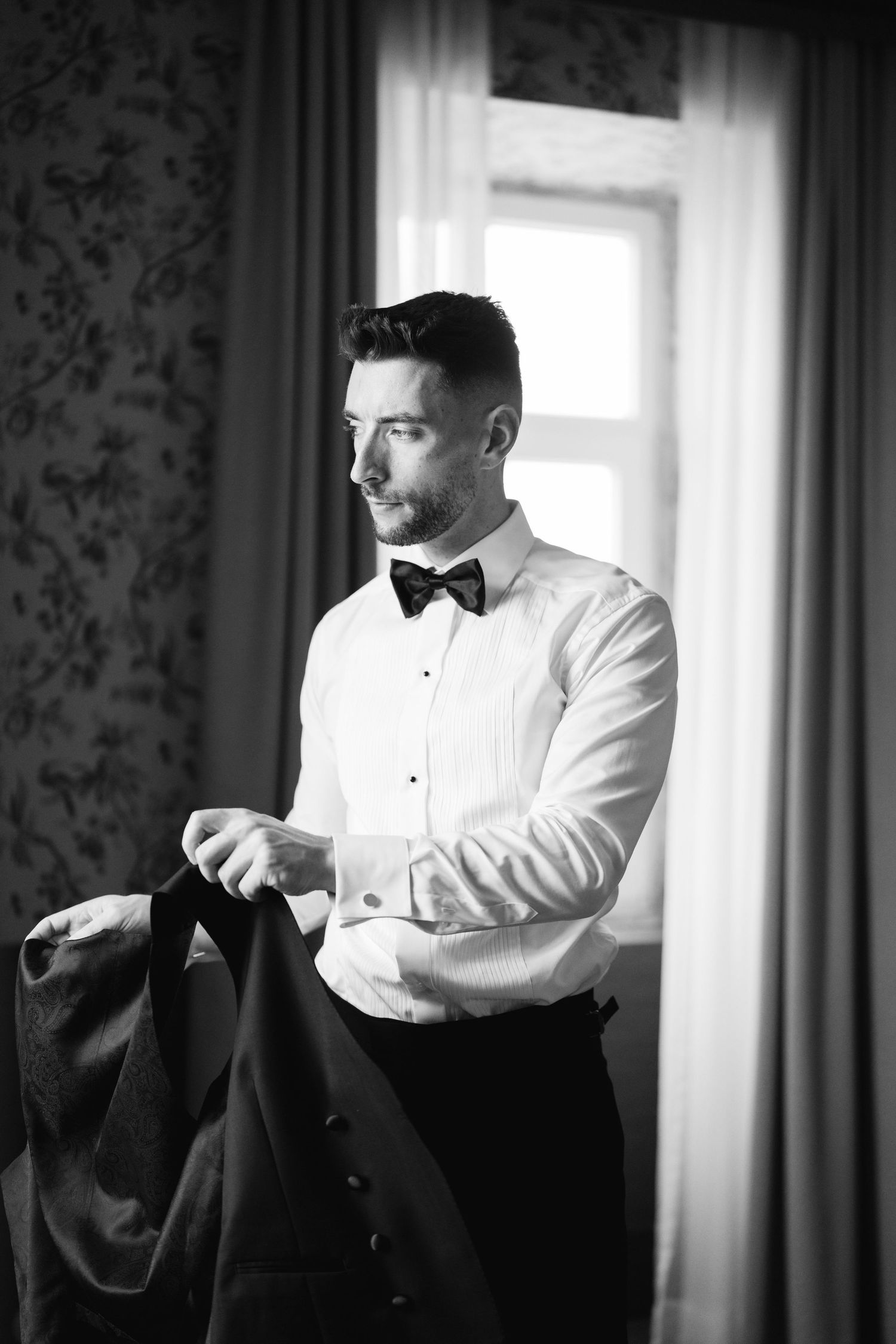 Black and white photo of someone putting on formal attire near a window.
