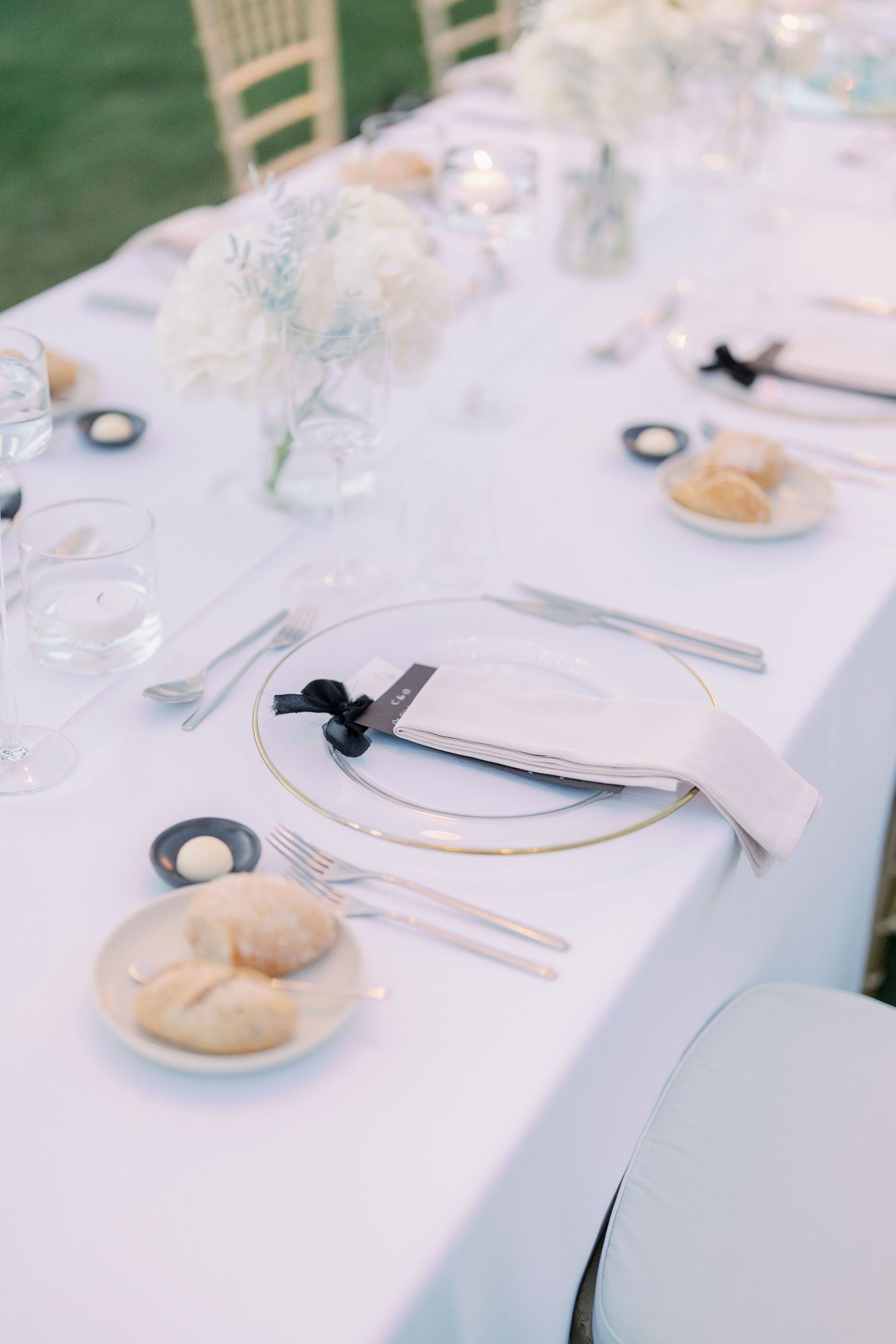 Minimalist white place settings with gold chargers at elegant outdoor wedding reception.