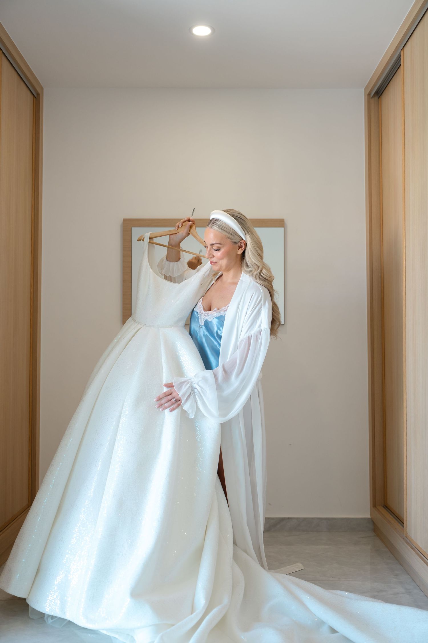 Bride in white robe admiring wedding dress hanging in dressing room.
