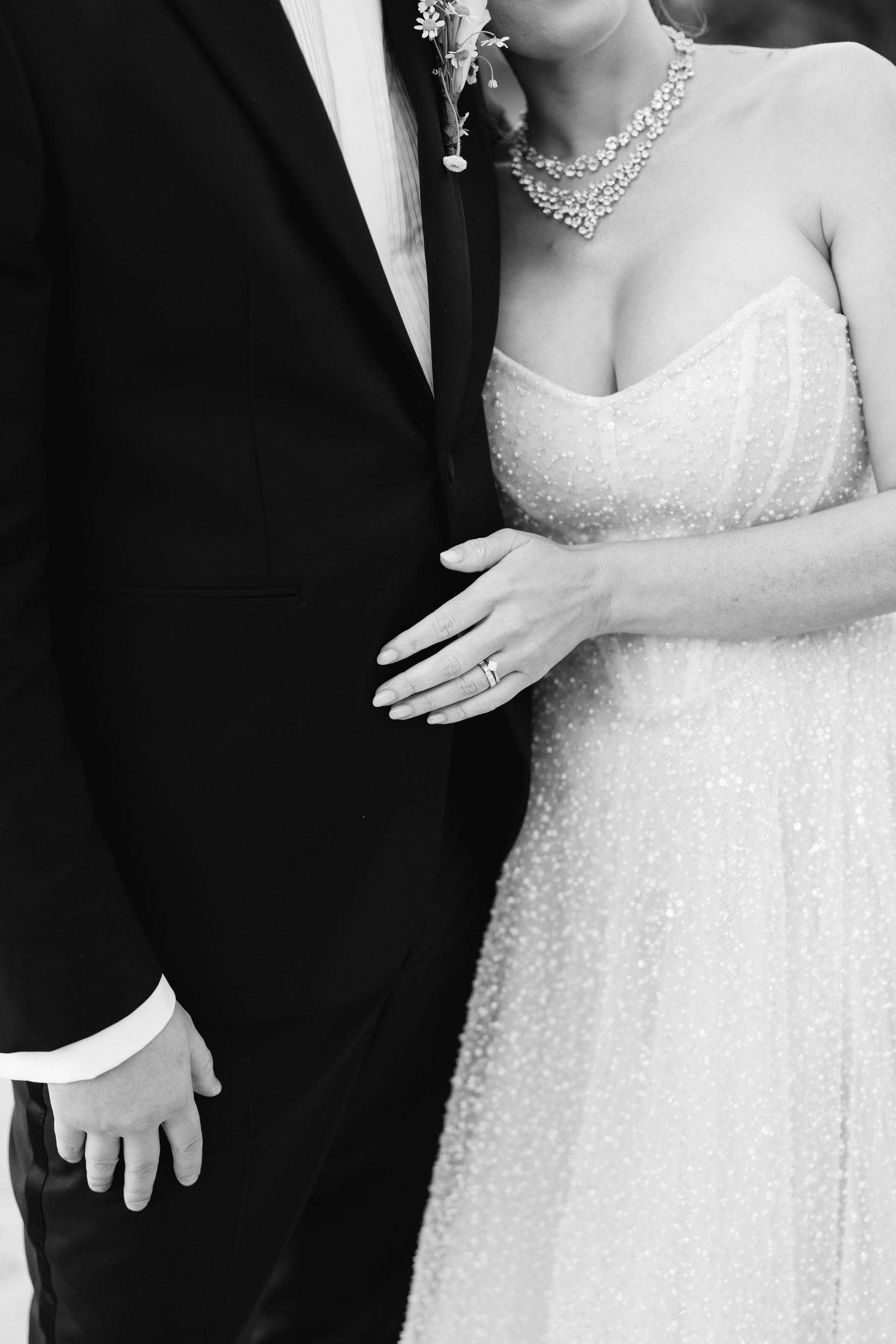 Black and white wedding photo showing bride in beaded gown with necklace standing close to groom in classic tuxedo.