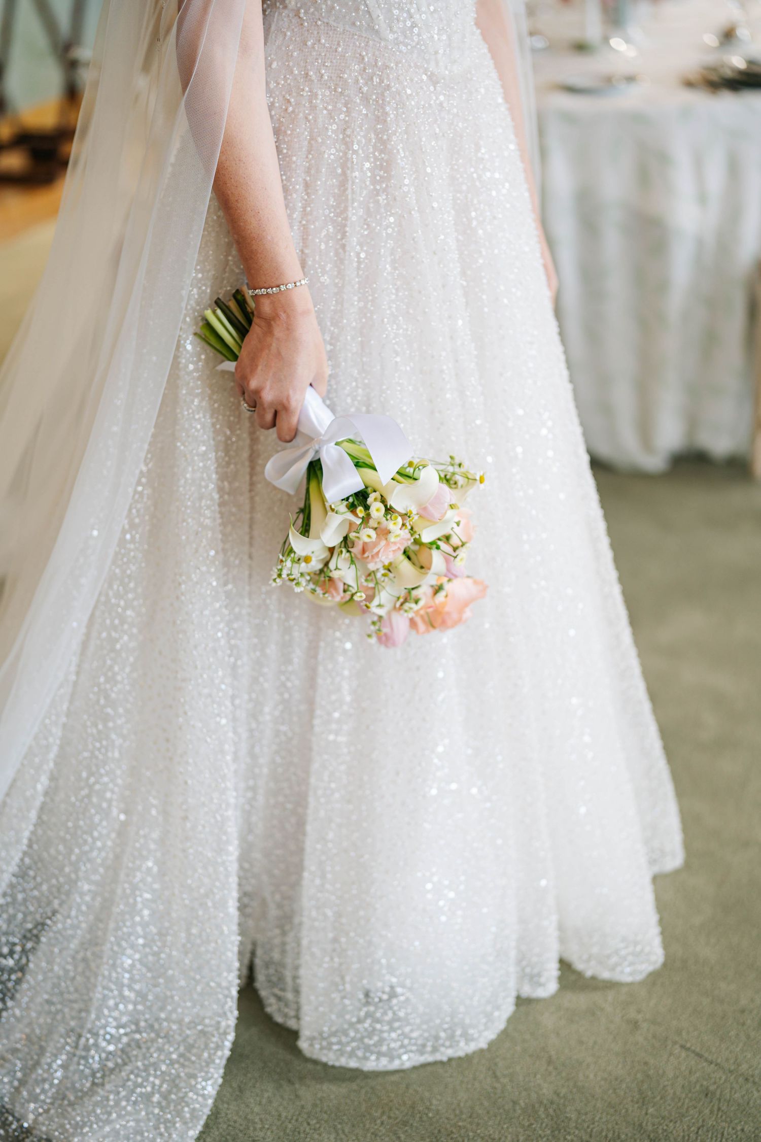 Close-up view of a sparkly white wedding dress with delicate tulle fabric and a small pastel floral bouquet.