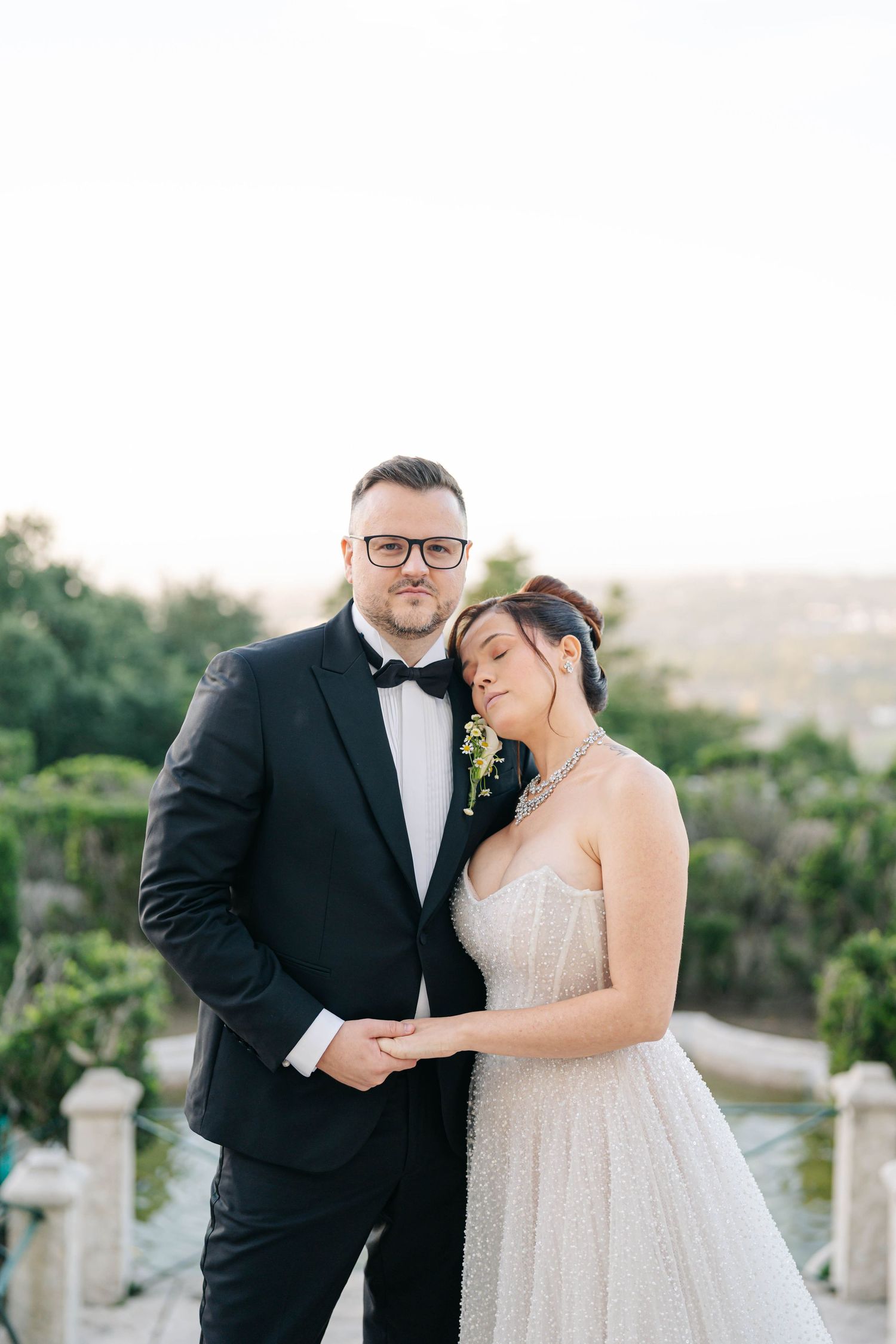 Elegant couple in formal wedding attire embrace on a scenic balcony with greenery in the background at sunset.