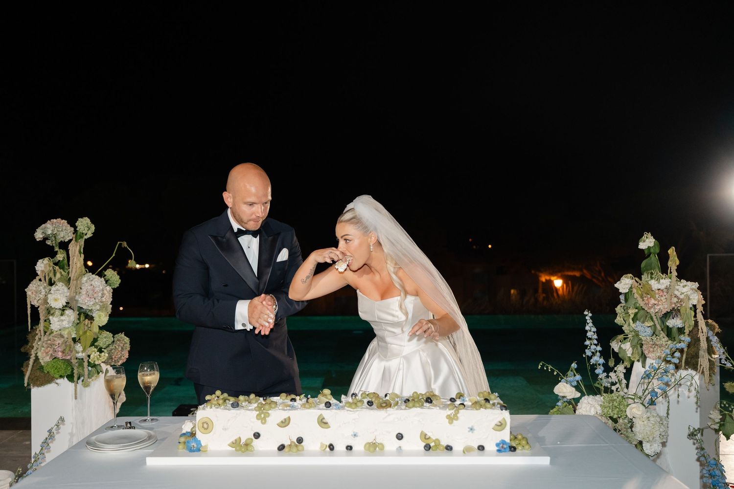 Couple cuts their elegant white wedding cake decorated with flowers at an evening reception.