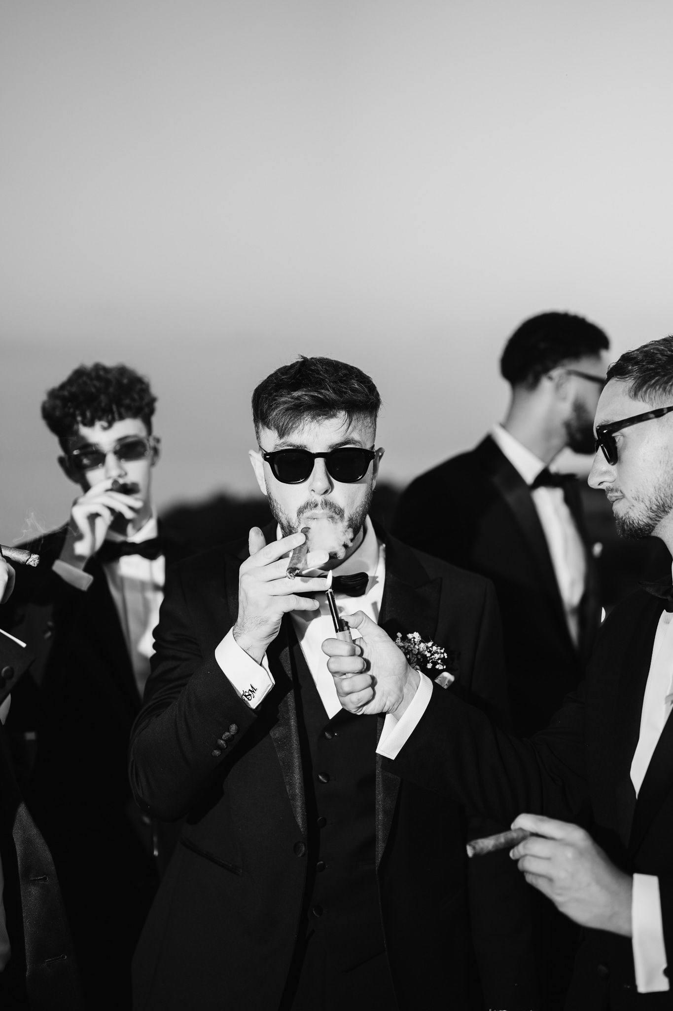 Wedding party members in black suits and sunglasses smoke together in artistic black and white.