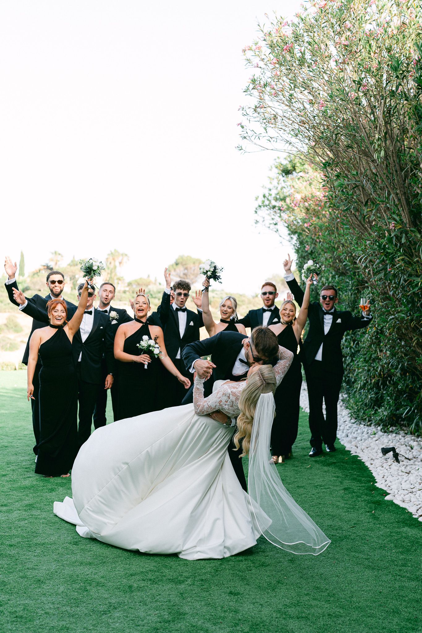 Wedding party in black and white attire strikes energetic poses on green lawn.
