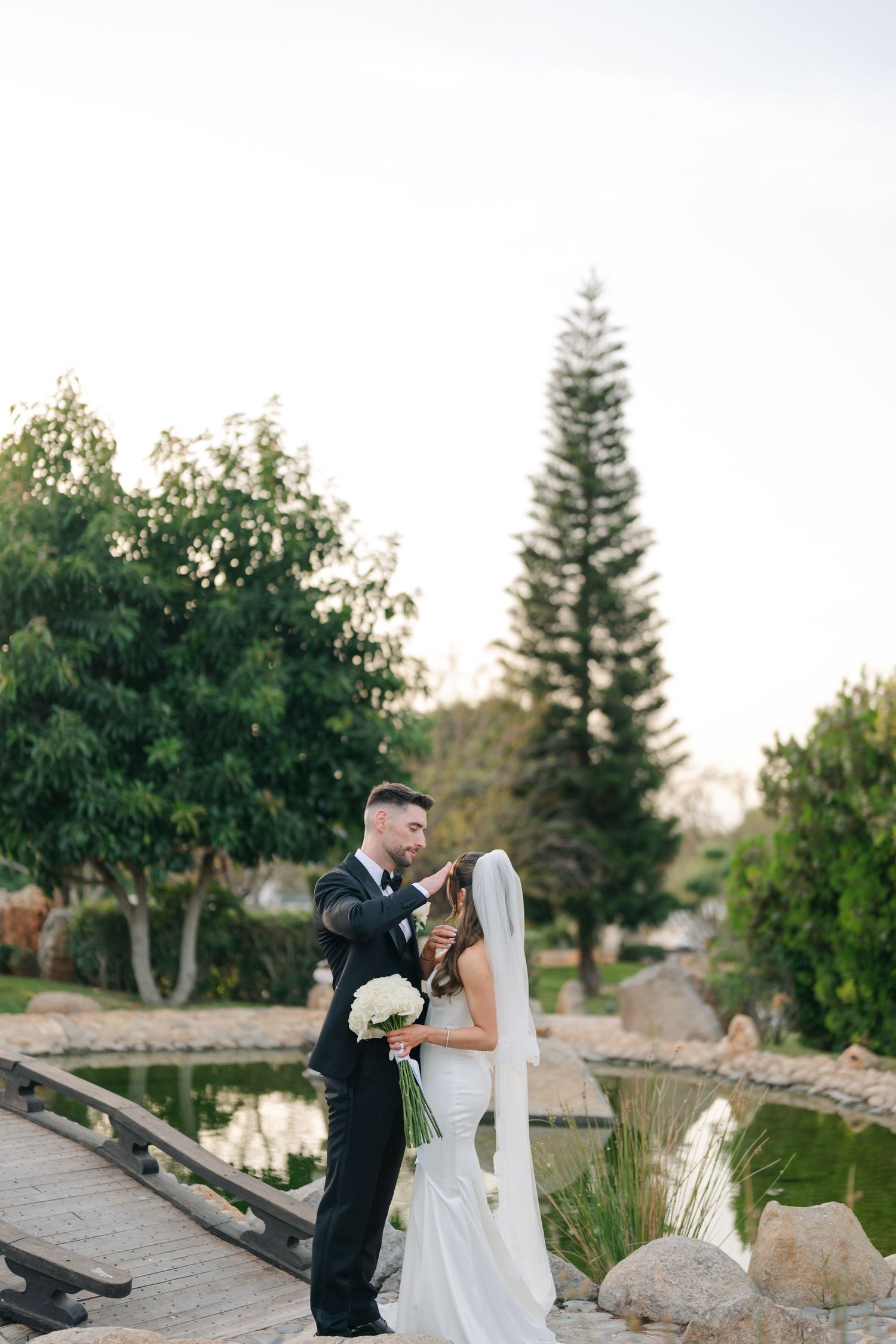 Wedding couple poses together near a tranquil pond with evergreen trees in the background.
