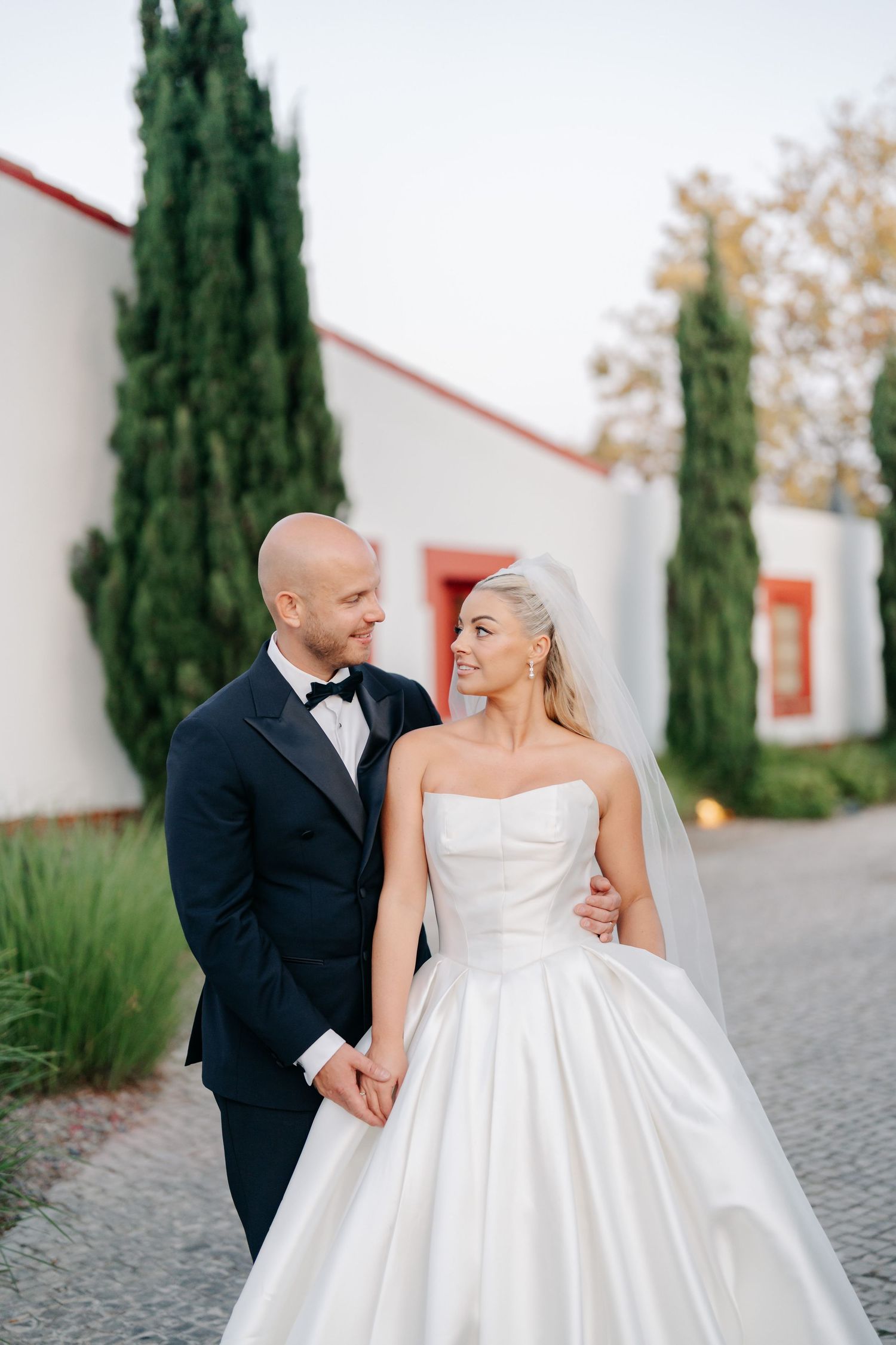Elegant couple in wedding attire walking together along cypress-lined path.