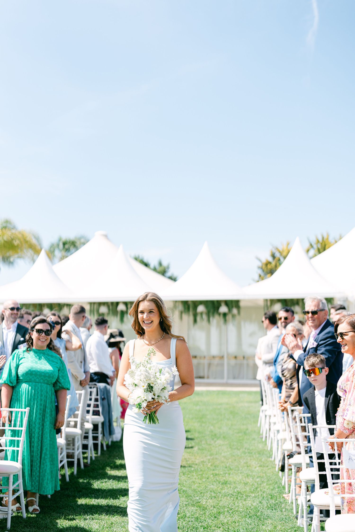Wedding attendees gather on a grassy lawn with white chairs and tents in the background.