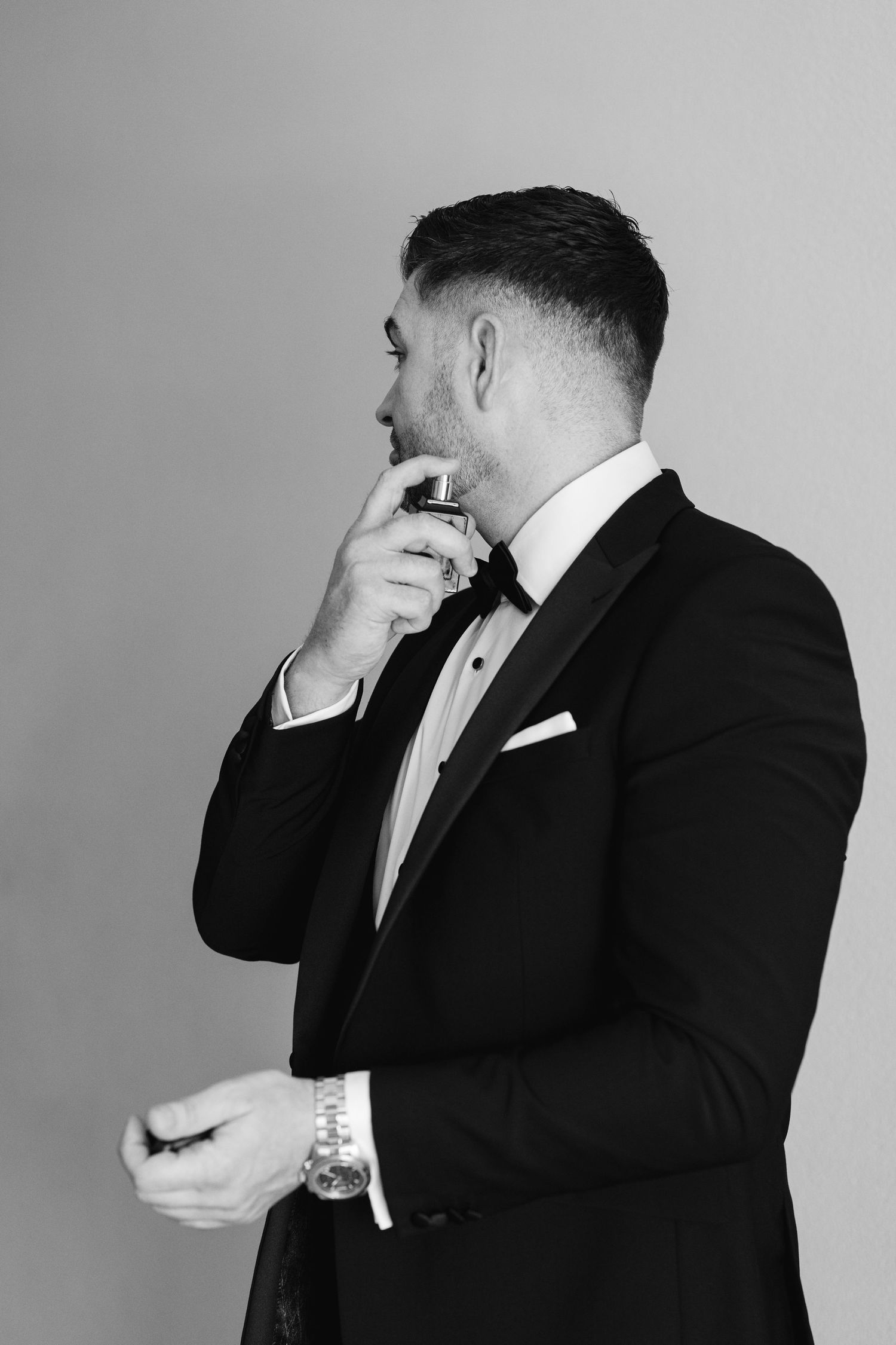 A groom in a black tuxedo and bow tie adjusts his collar in a black and white portrait.