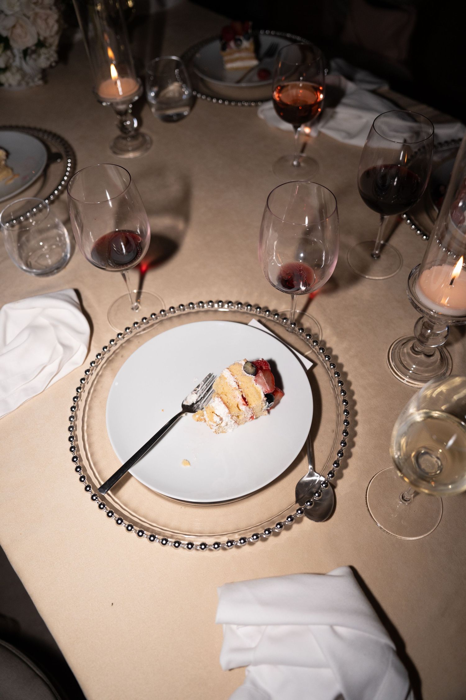 Elegant table setting with wine glasses and a slice of cake on a beaded charger plate illuminated by candlelight.