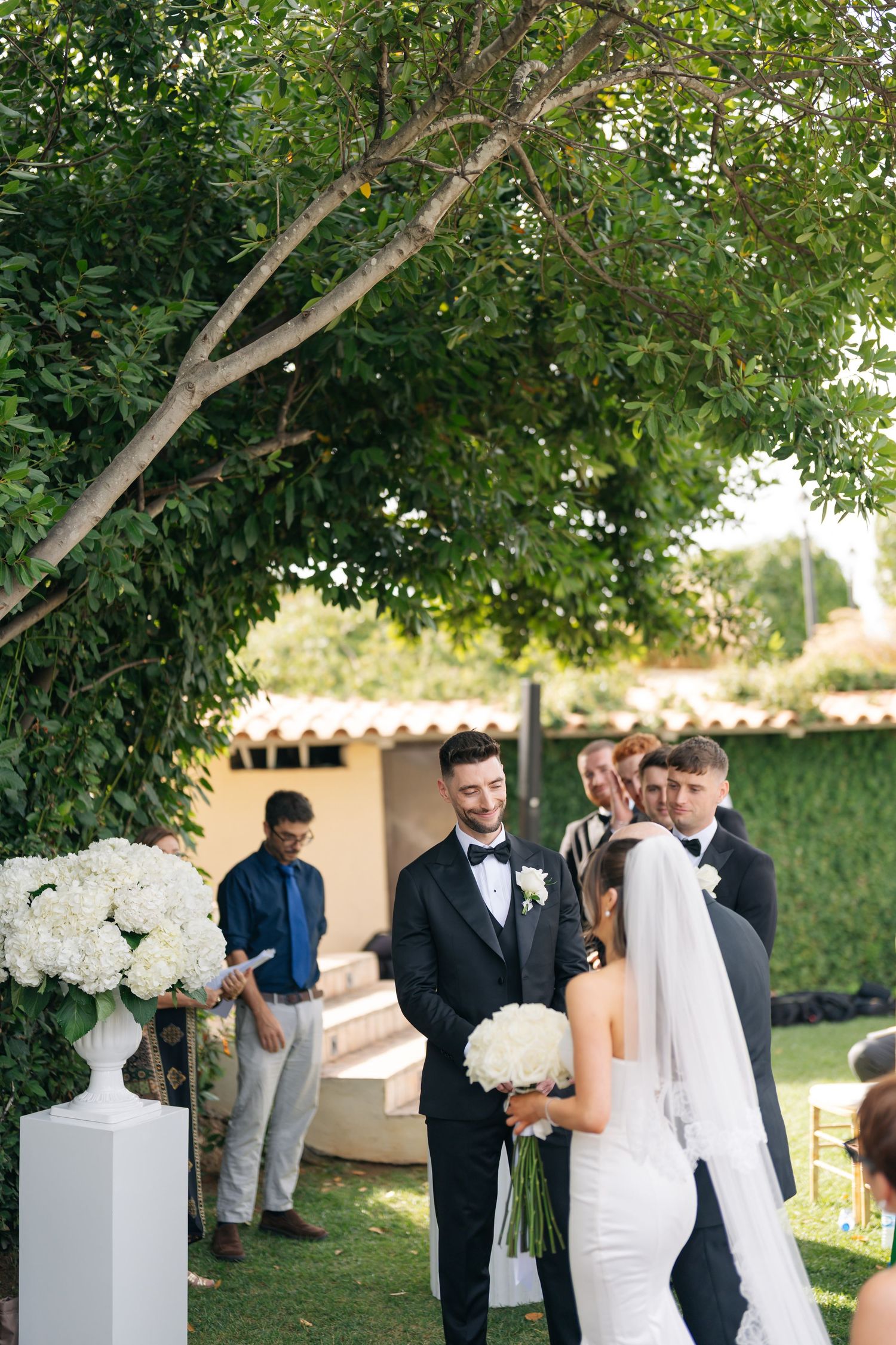 Wedding party members gather under trees near white floral arrangements at an outdoor ceremony.