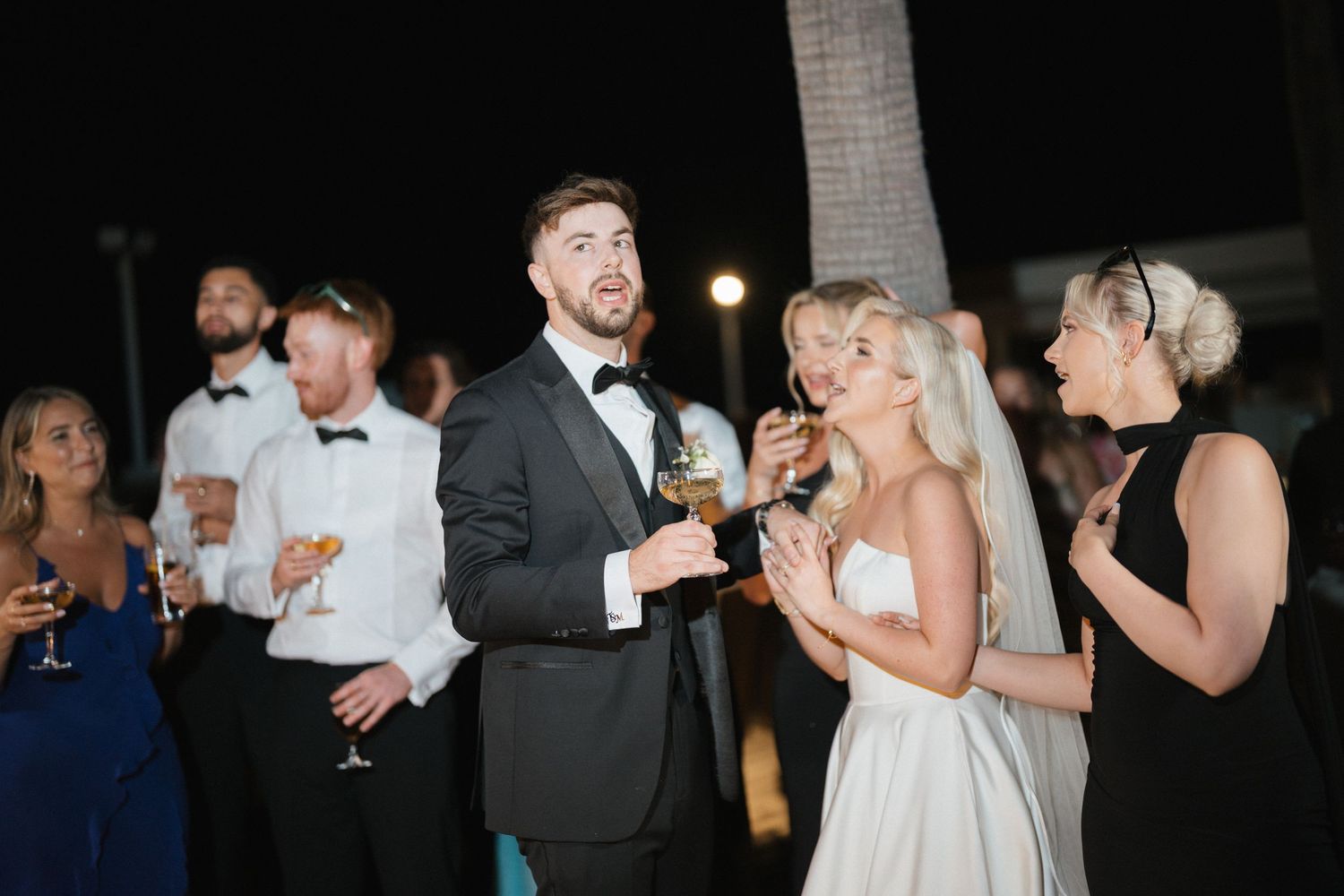 Wedding celebration scene with guests in formal attire enjoying drinks and laughter at an evening reception.