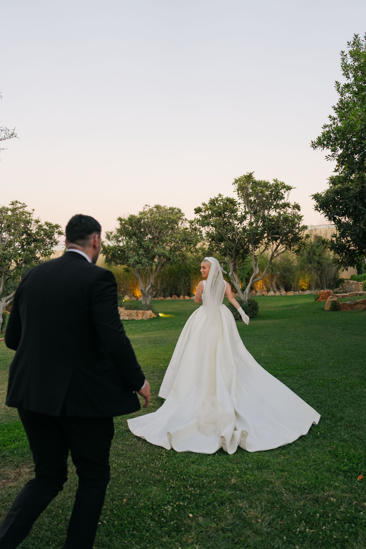 Wedding couple walking across a green lawn at sunset with flowing white dress.