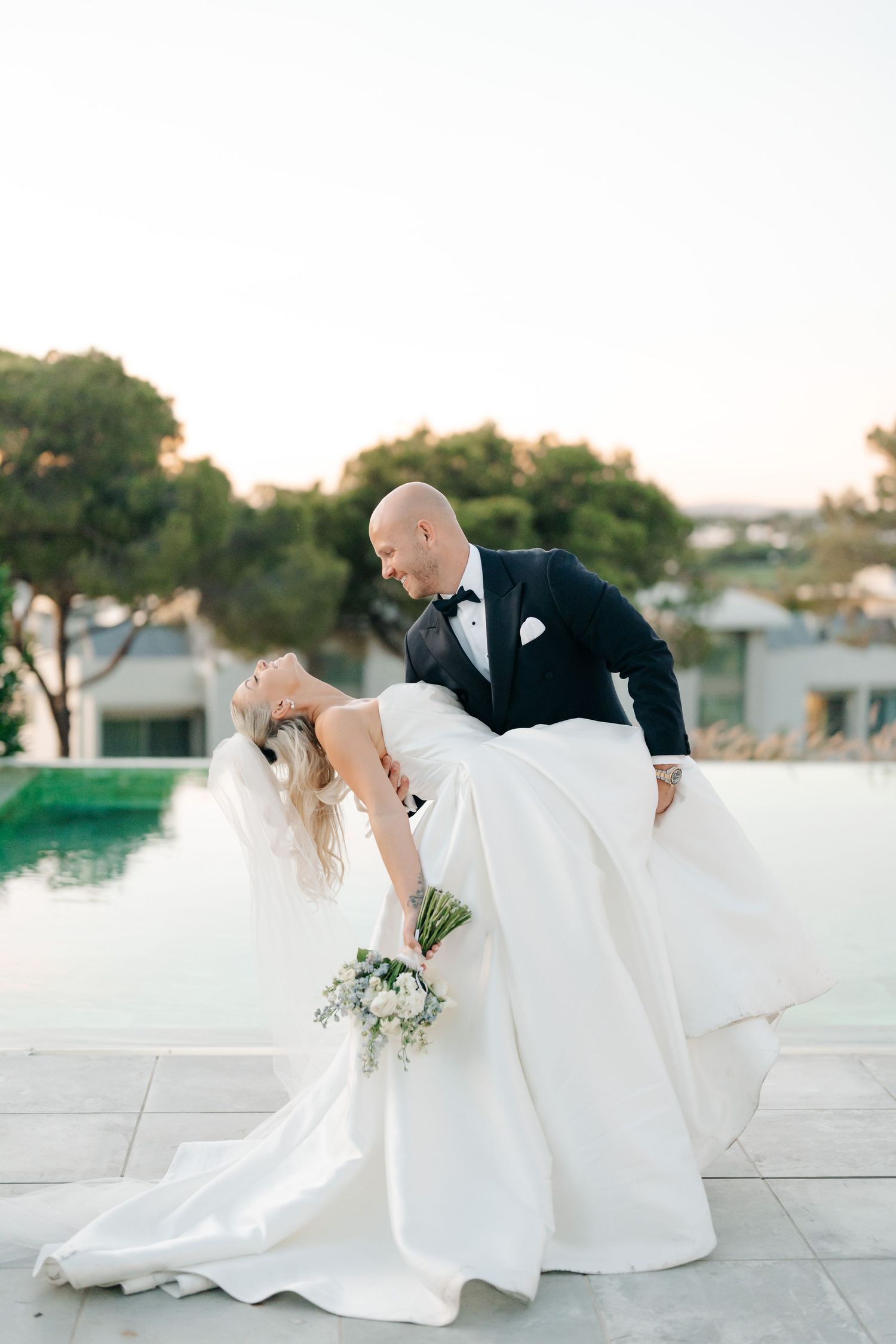 Dramatic wedding pose with flowing white dress against sunset poolside setting.
