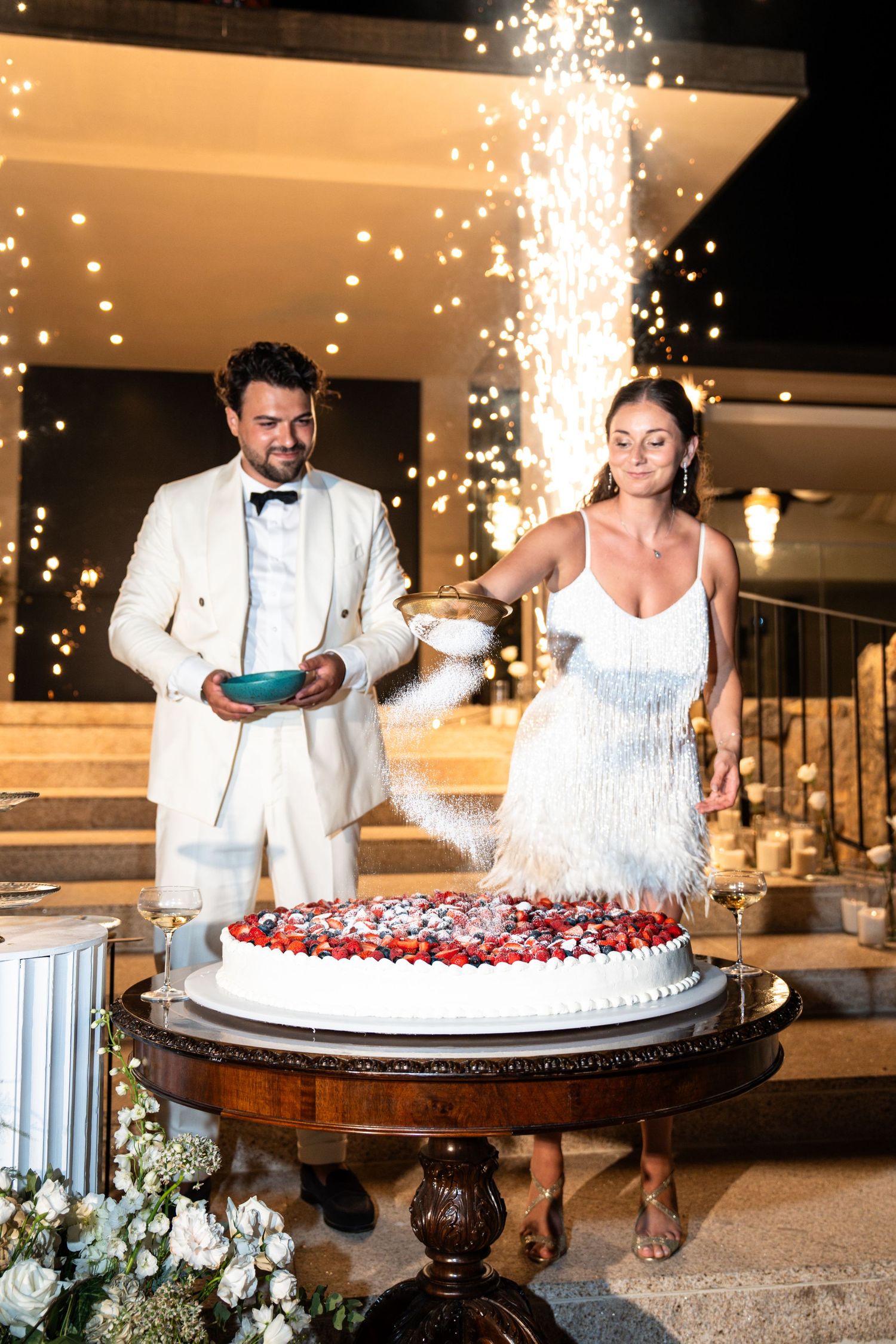 Couple in white attire cutting a large wedding cake with sparklers creating a magical effect at their reception.
