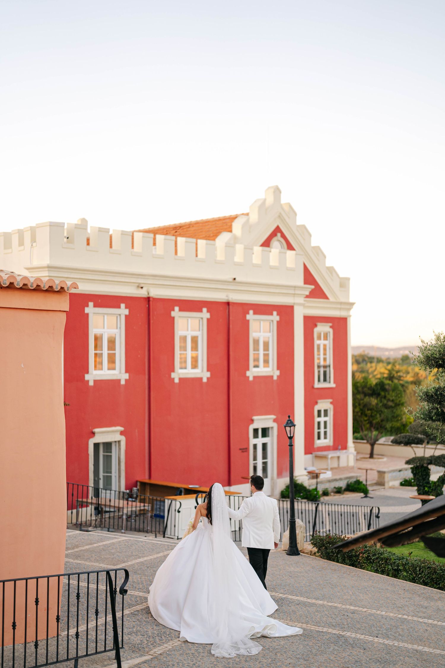 A couple in wedding attire stands outside a pink Mediterranean-style building with white trim and orange roof tiles.