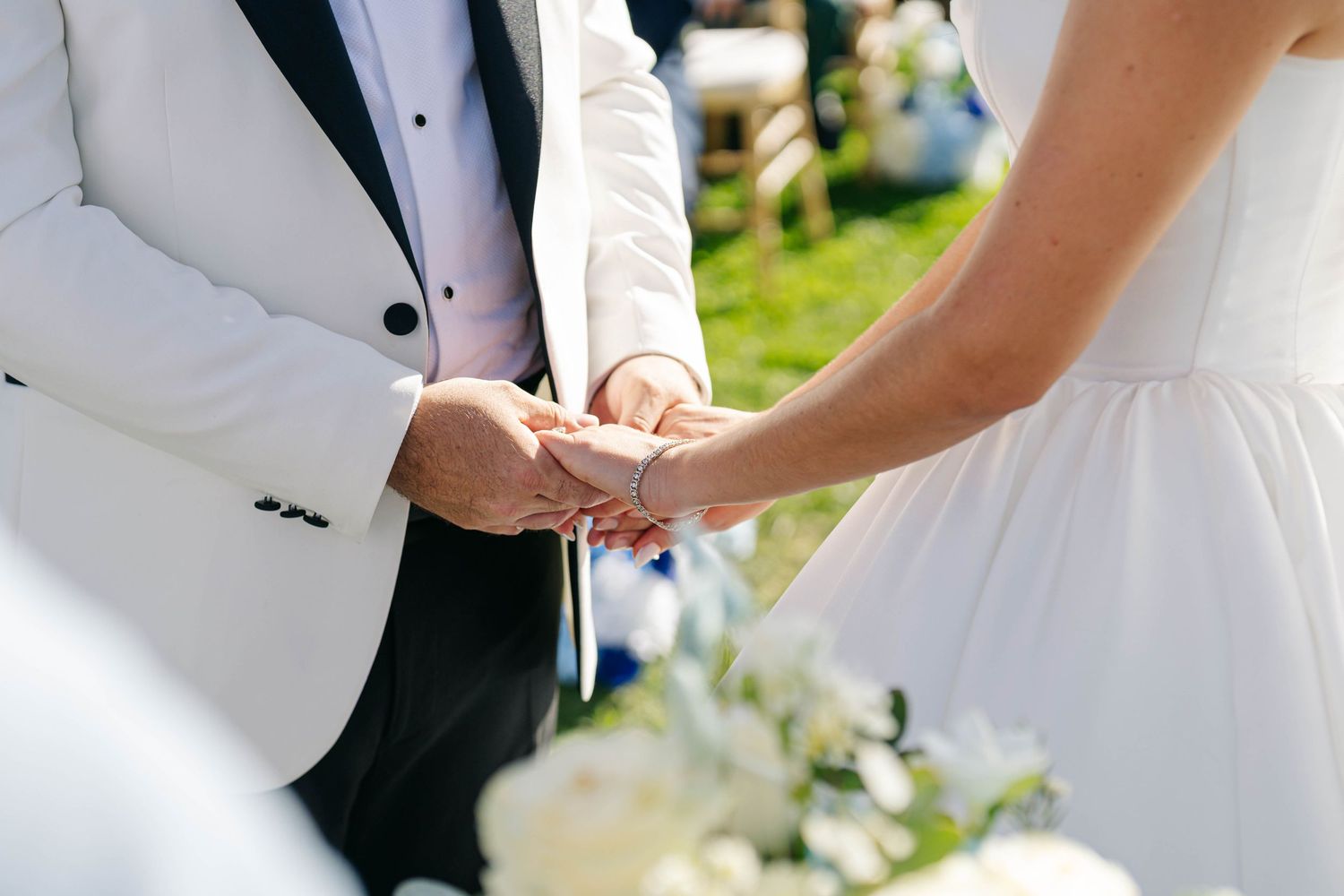 Bride and groom hold hands during outdoor wedding ceremony with guests seated in background.