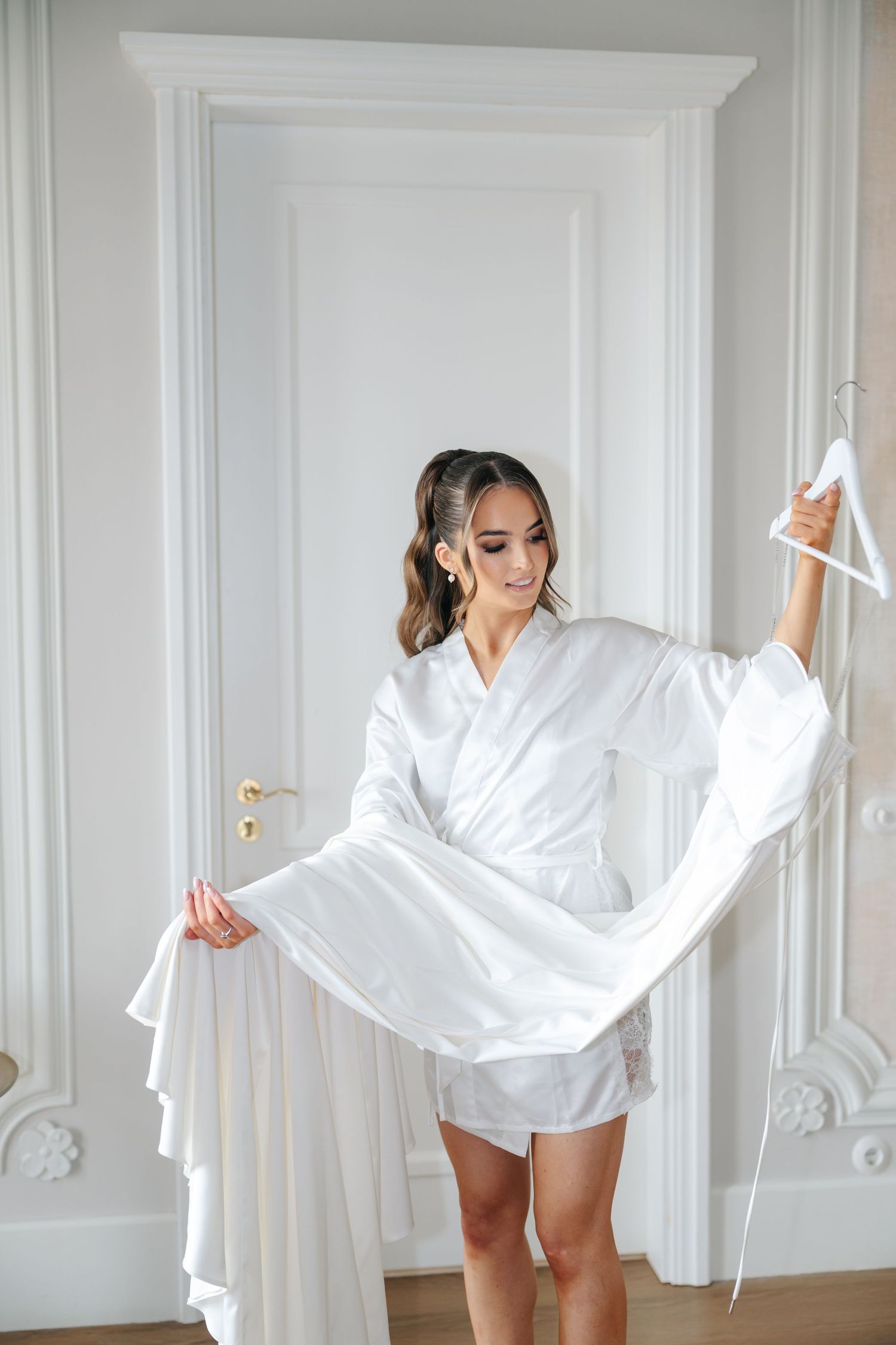 Woman in white robe holding wedding dress in bright room with ornate moldings.