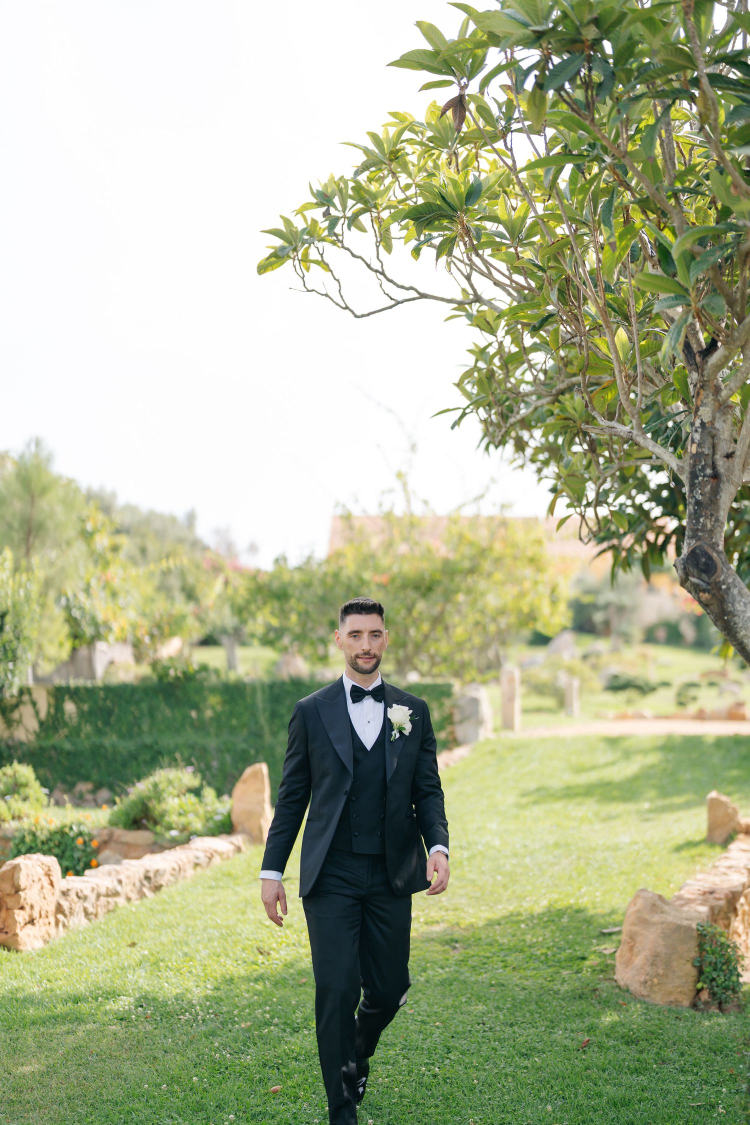 A groom in formal attire walks along a grassy path with trees in the background.