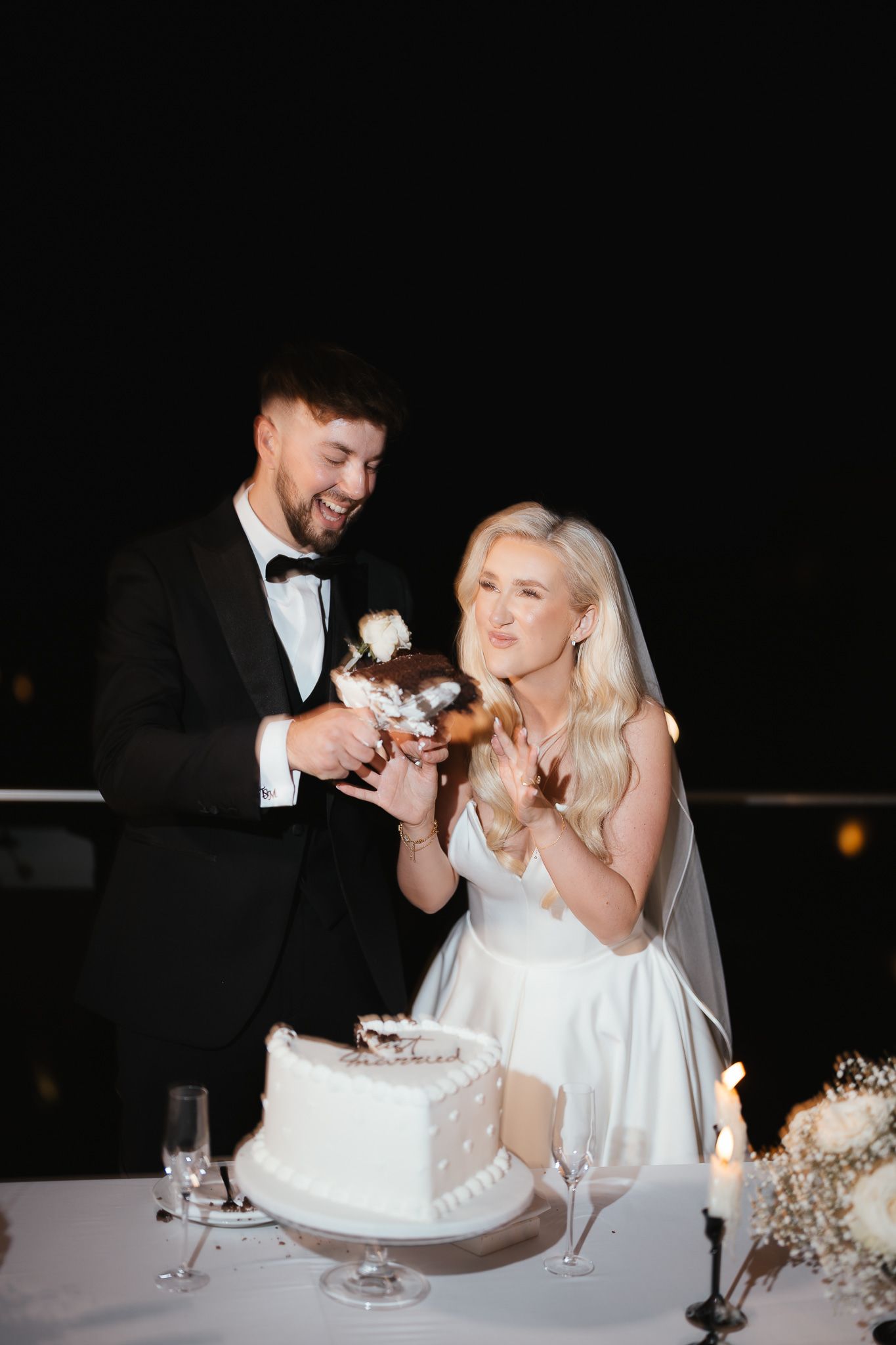 Newlyweds laugh while sharing wedding cake at candlelit reception.