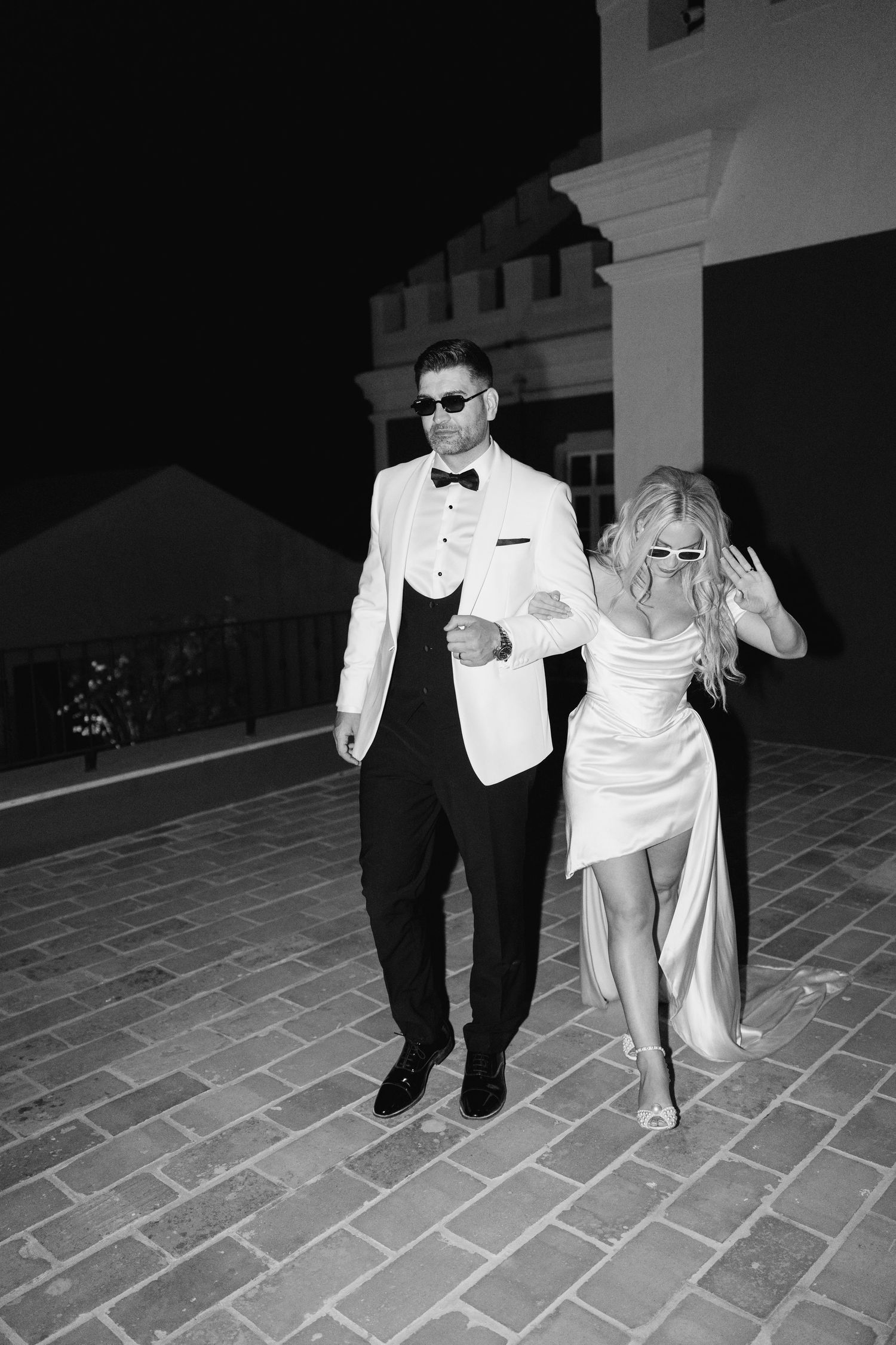 A couple in formal attire walking together on brick pavement at night.