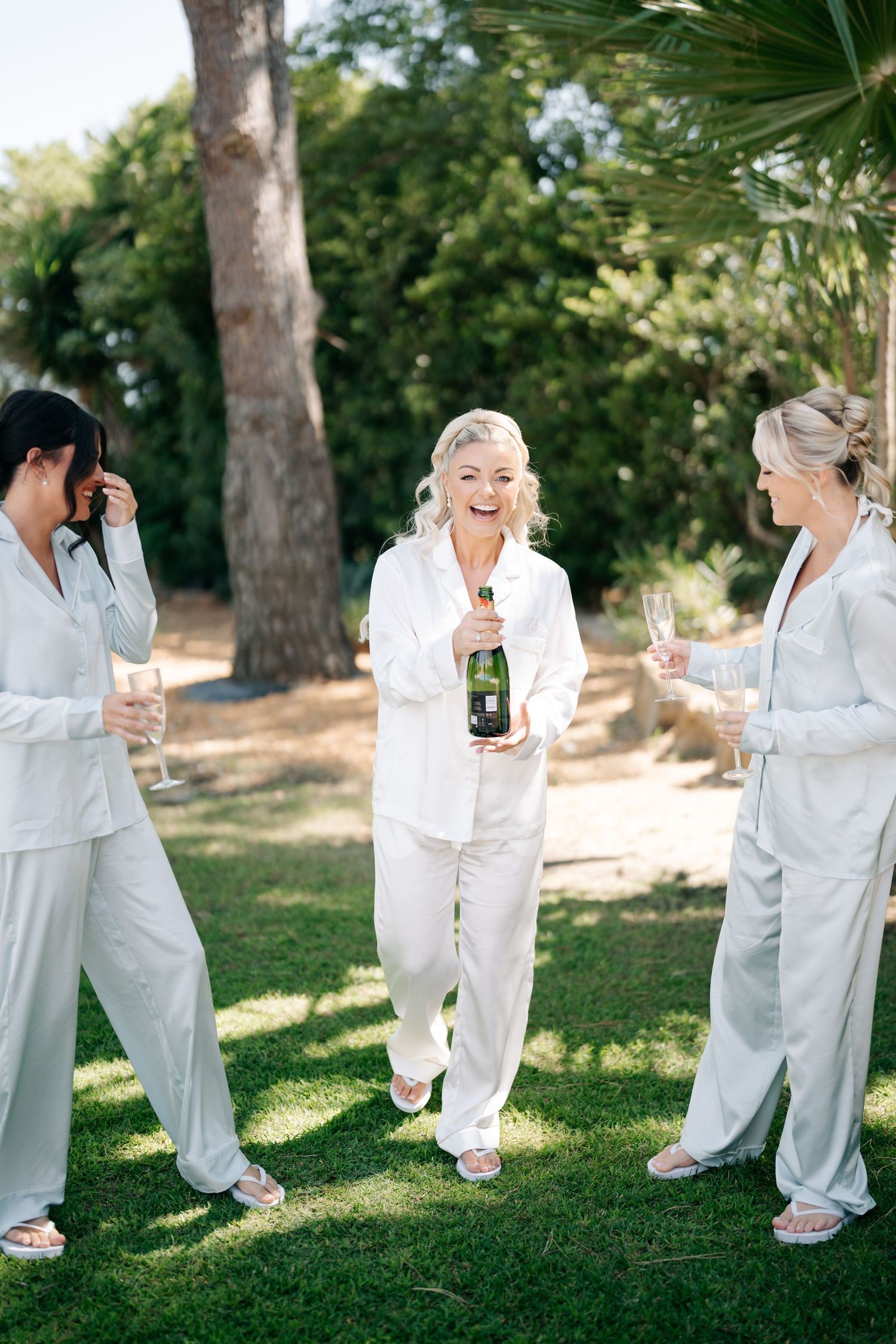 Group wearing white loungewear laughs together while holding champagne glasses outdoors.