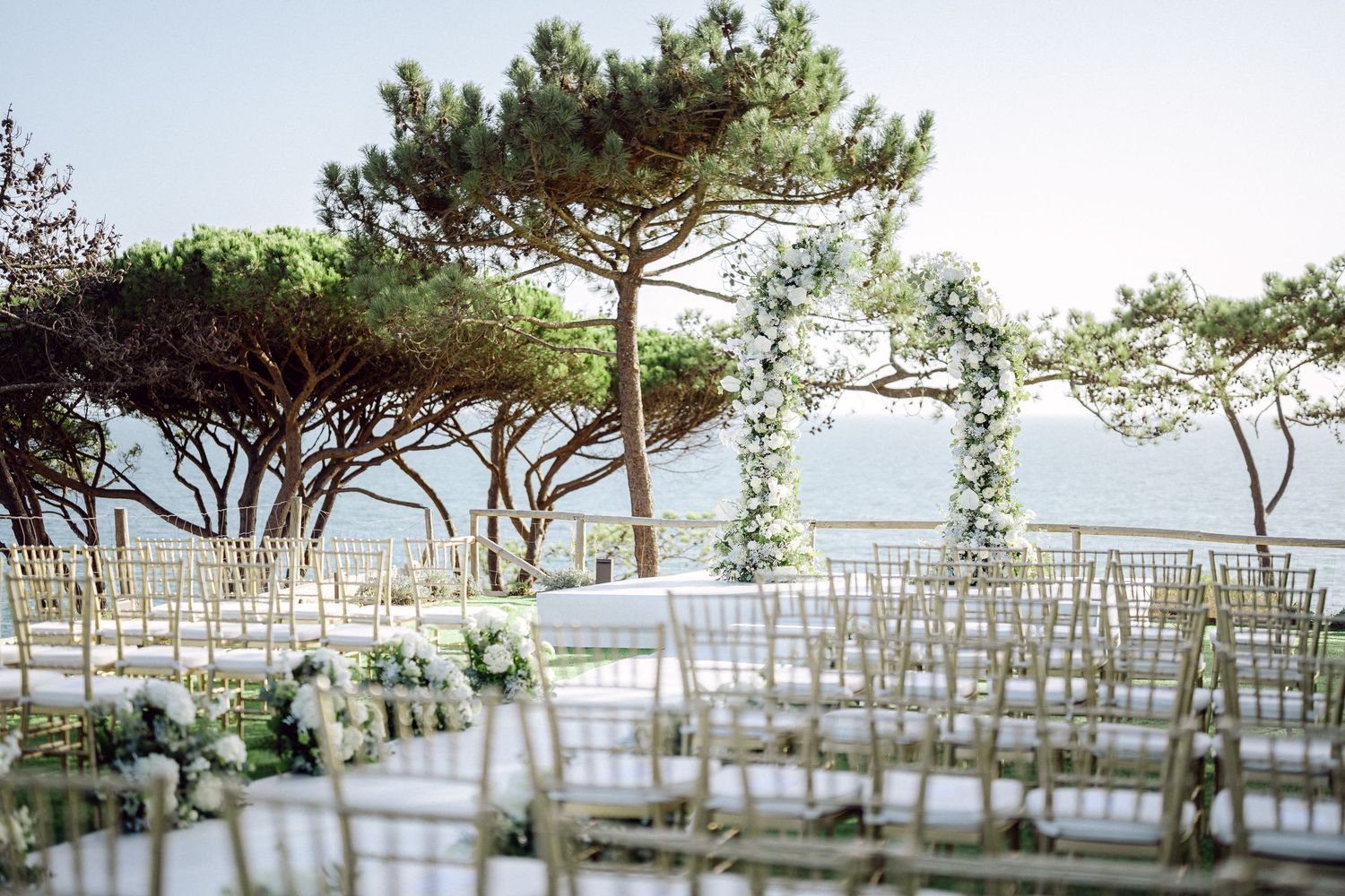 Elegant outdoor wedding setup with white flowers and gold chairs overlooking a serene waterfront.