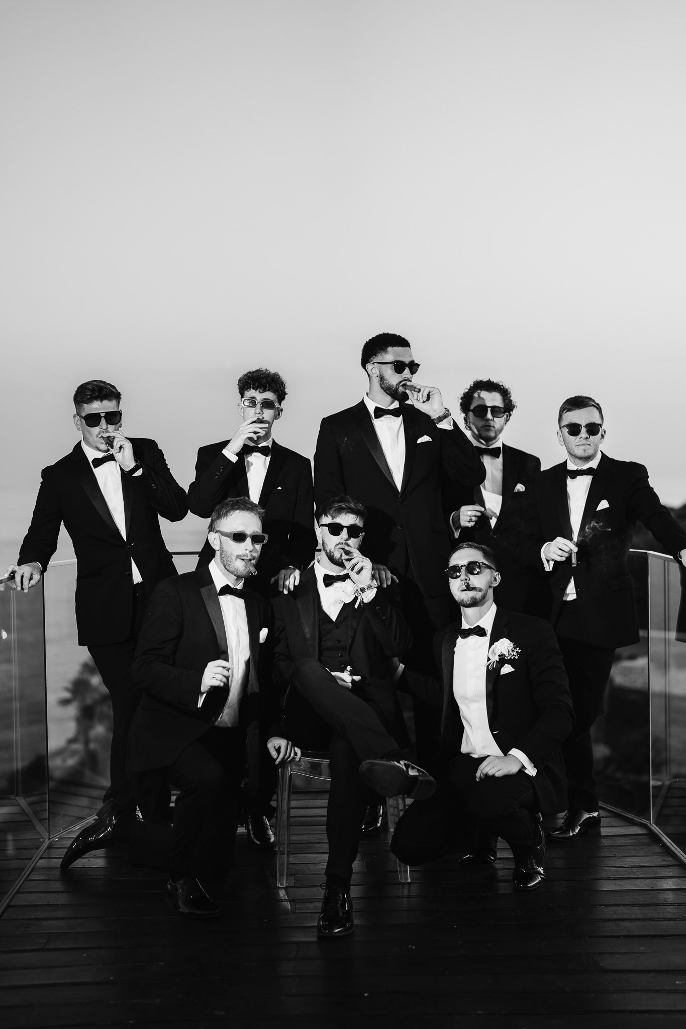 Groomsmen in black tuxedos and sunglasses pose together in a stylish black and white photo.