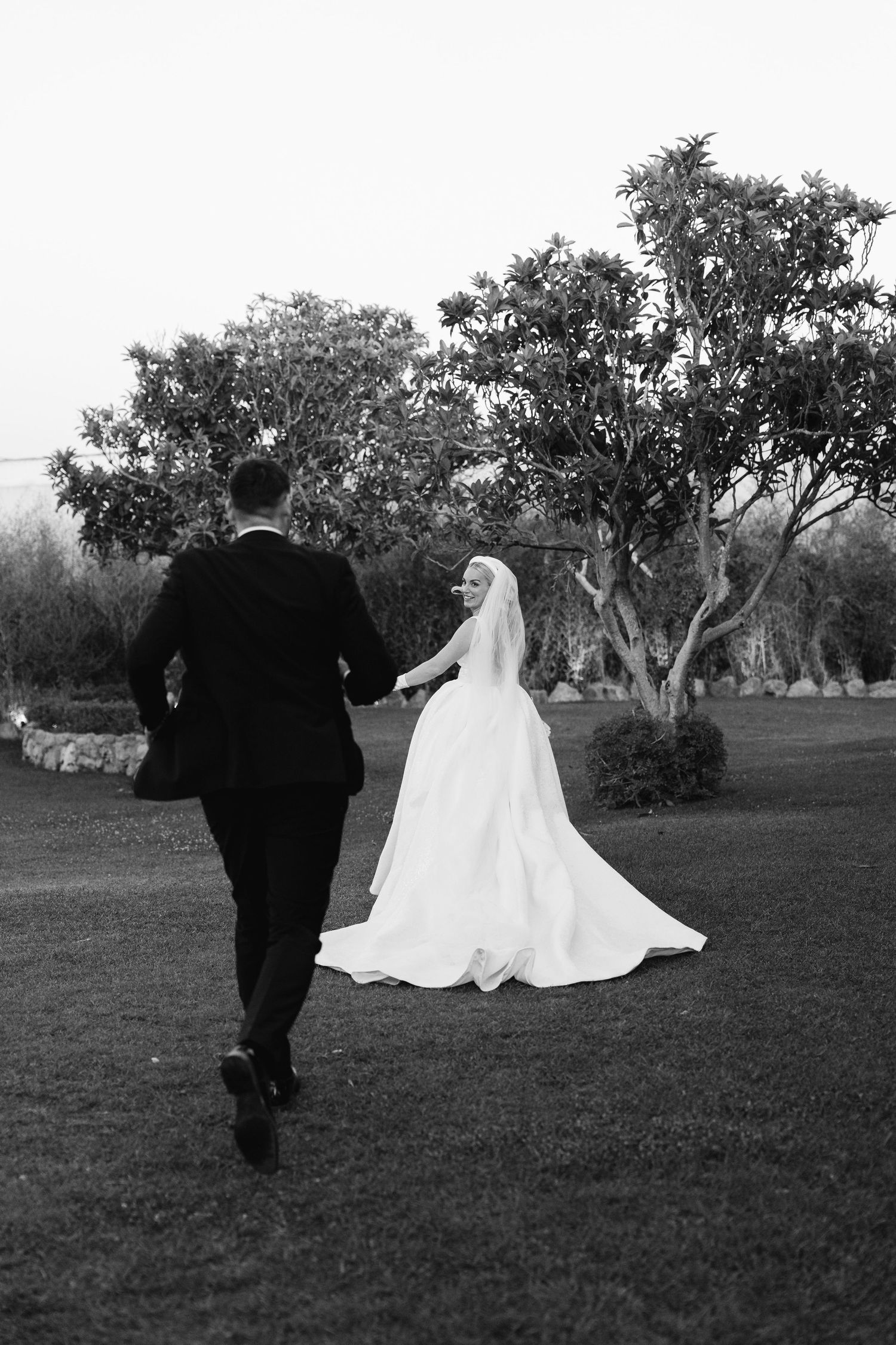 A bride in a flowing white dress and groom walk together on a grassy field at dusk.