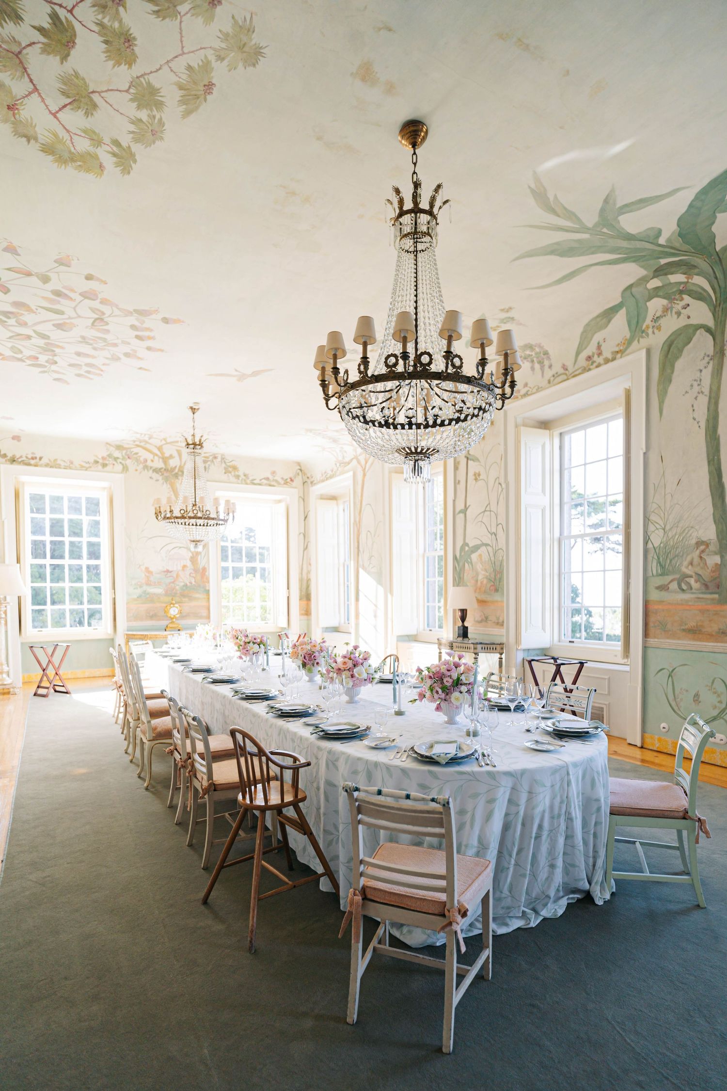 Elegant dining room with white tablecloths, ornate chandeliers, and delicate floral wall murals set for a formal event.