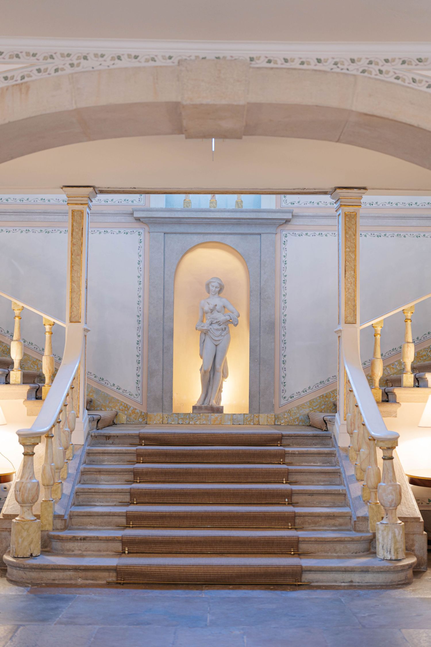 Classical interior with stone steps leading to arched alcoves containing statues, illuminated by warm lighting.