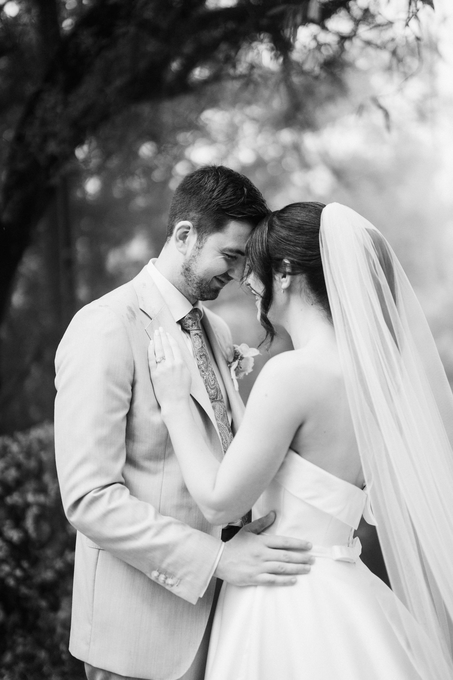 A tender black and white wedding moment captured outdoors with the bride in white and groom in light suit embracing.